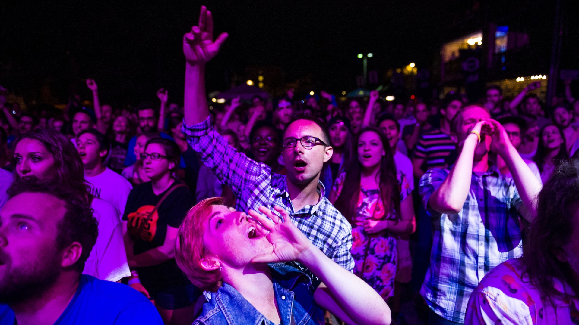 A woman in the foreground with her head titled backward and hands on her mouth yells while a man stands behind her, with his hand in the air. They are surrounded by people showered in pink and purple lighting as they face a musical stage. 