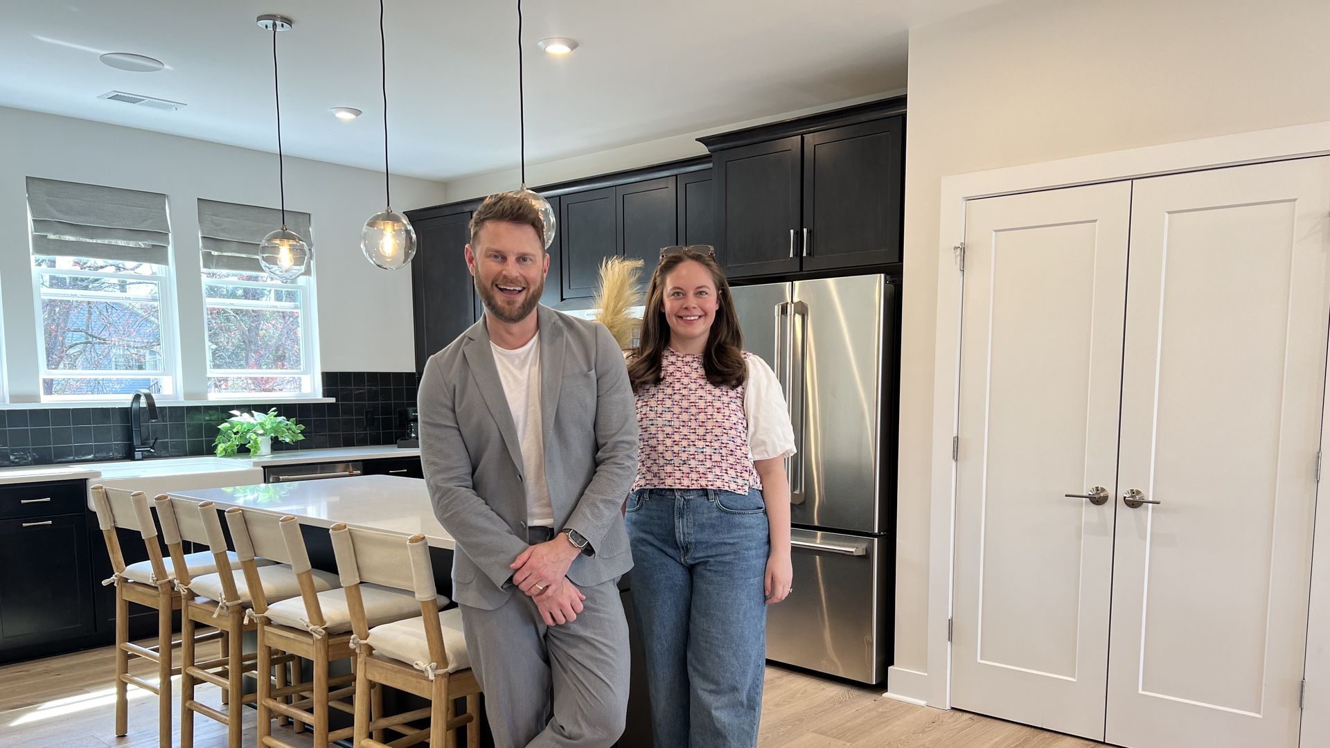 designer bobby berk in a gray suit standing in a kitchen with a reporter in a pink top and jeans