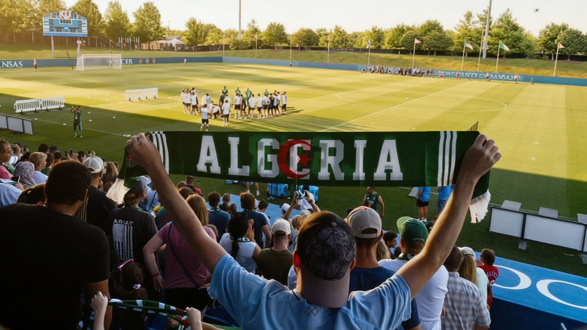 Sunlit soccer stadium with a team huddle on the striped green field. In the foreground fans hold a green scarf reading ALGERIA with a red crescent. A blue scoreboard and tall floodlights loom.