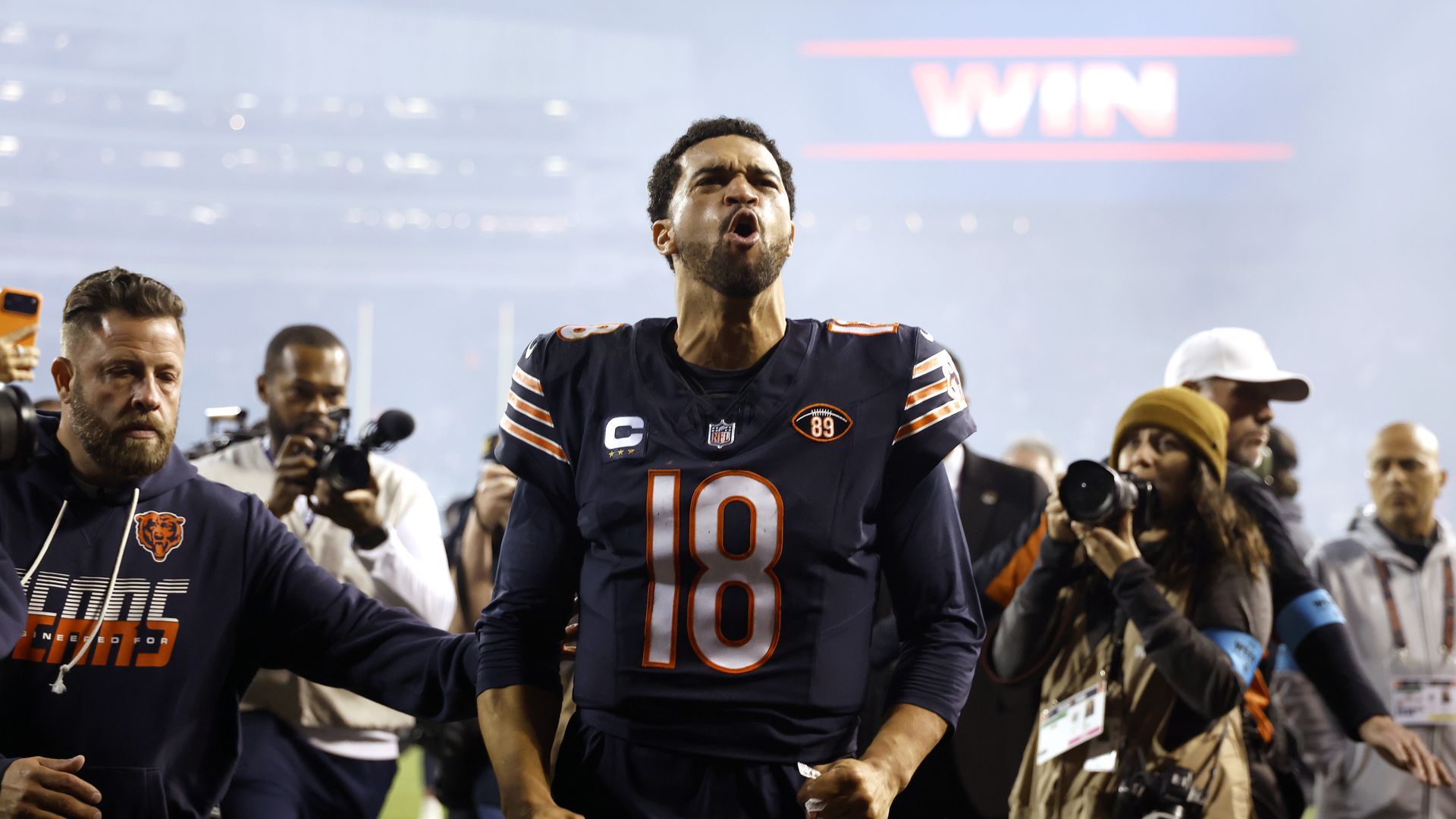 Chicago Bears player in a navy jersey with number 18 and a captain patch celebrates on the field, fists clenched, as reporters and fans surround him; a bright red WIN sign glows in the background.