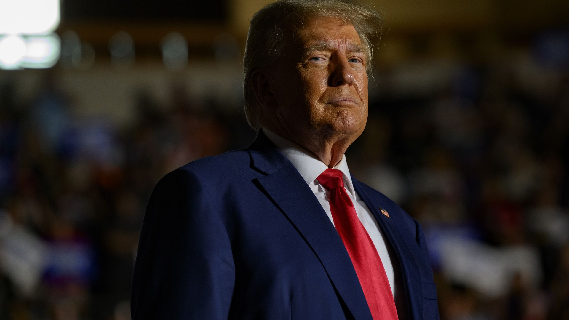 Donald Trump, wearing a blue suit and red tie, looks at the camera during a political rally.