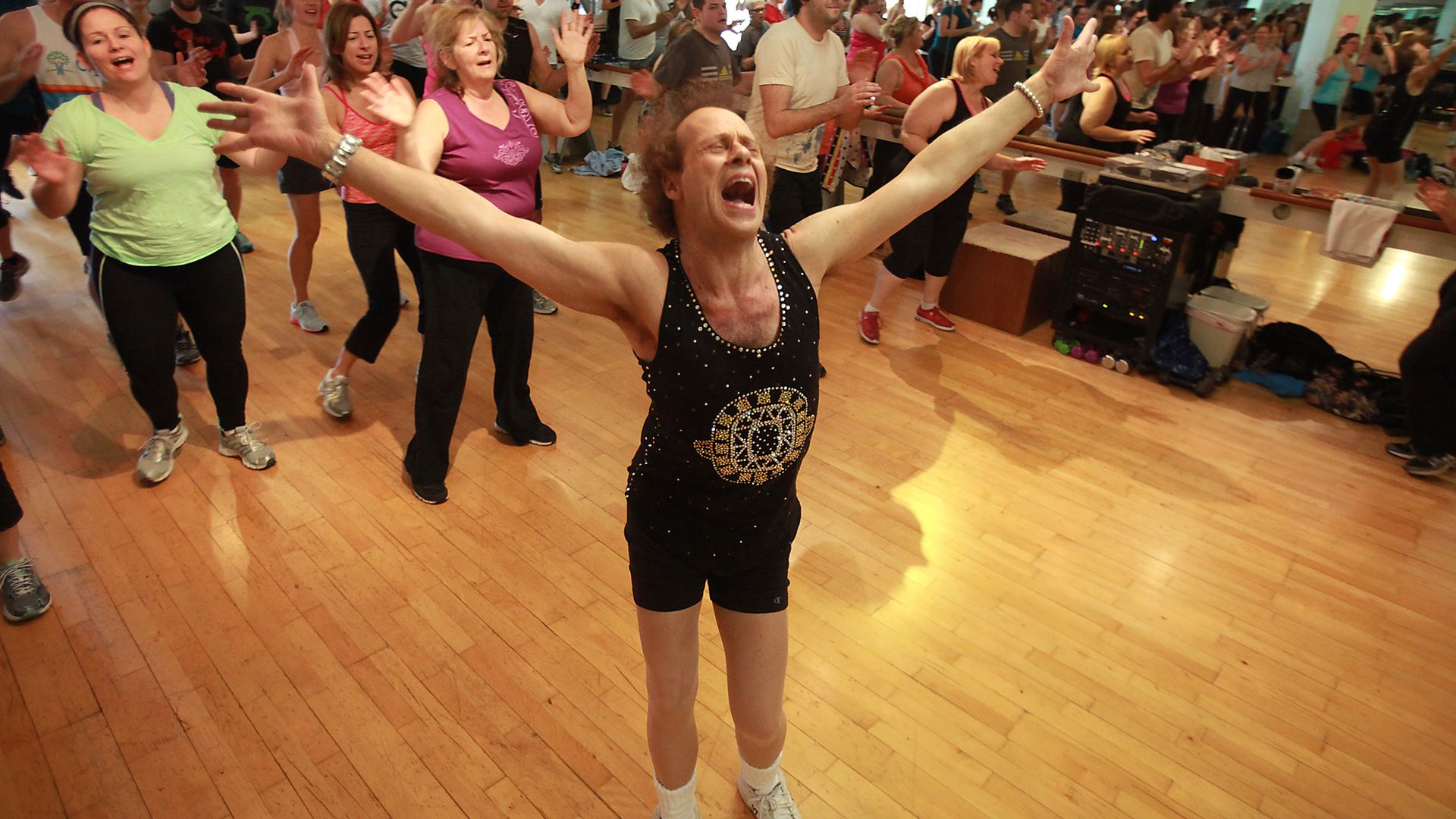 Richard Simmons smiles with his arms wide at the front of a busy workout class.