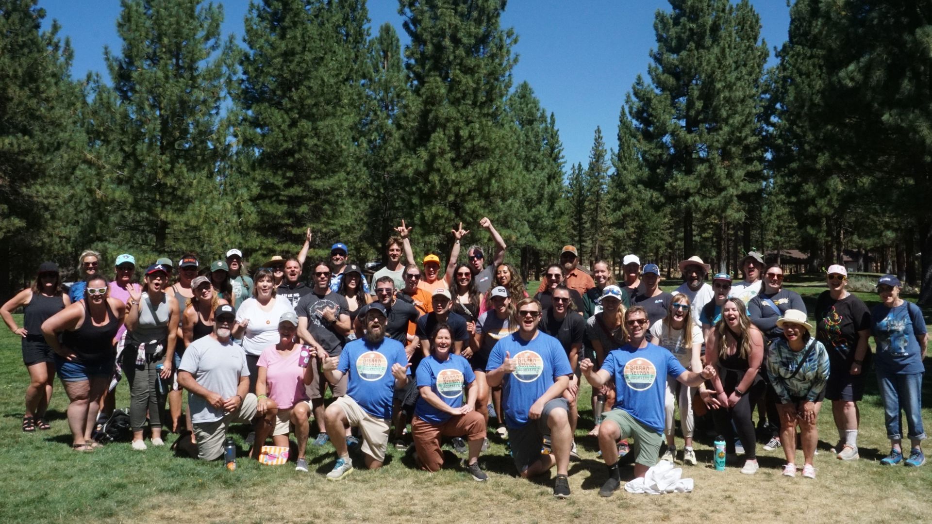 Large outdoor group photo in a park with tall evergreen trees and a blue sky; dozens of people wearing blue shirts and hats, smiling and posing together in front of the trees.