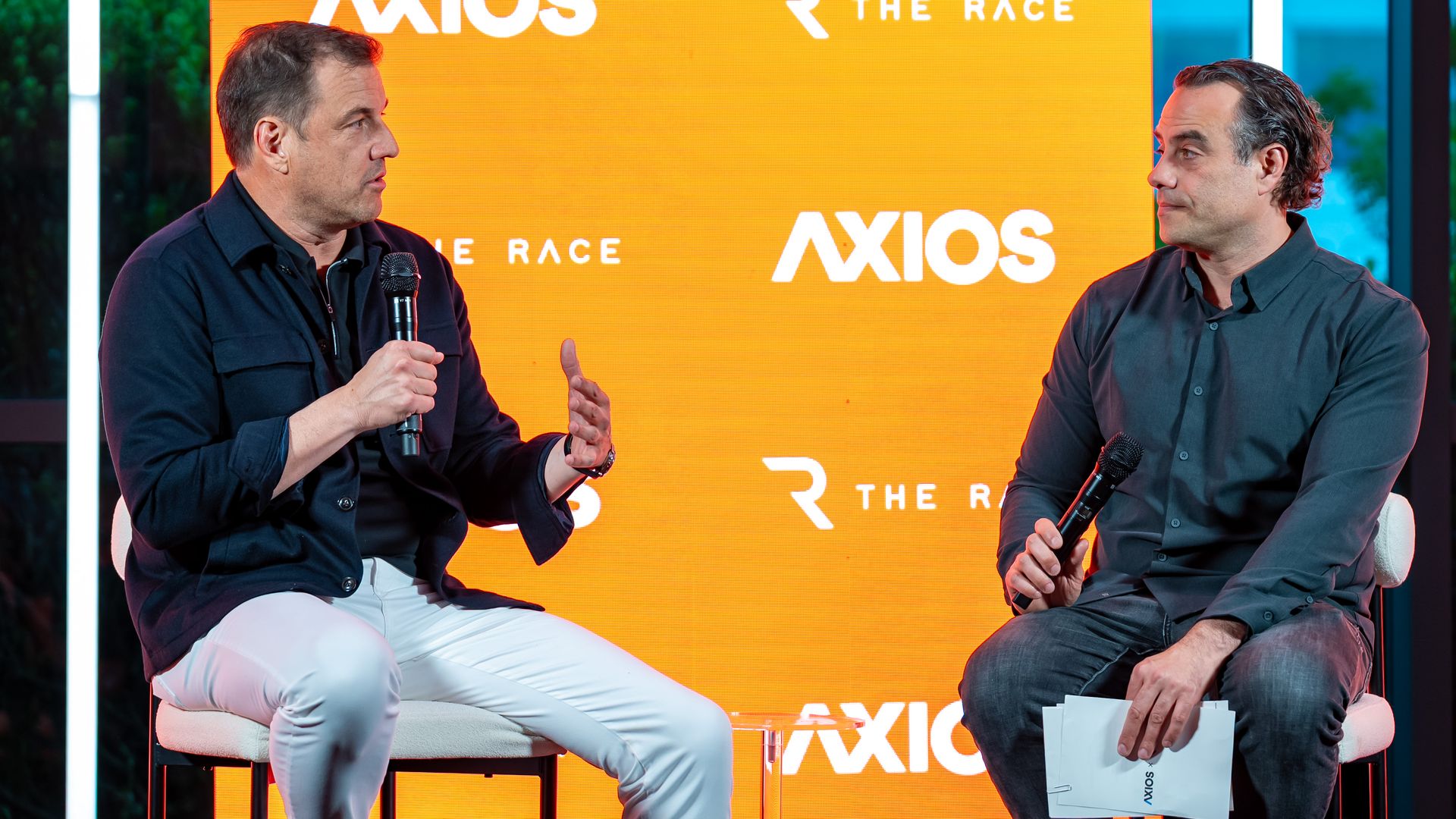 Two men sit on a stage in dark shirts, one holding a microphone and speaking while the other listens; orange backdrop displays white logos for "AXIOS" and "THE RACE".