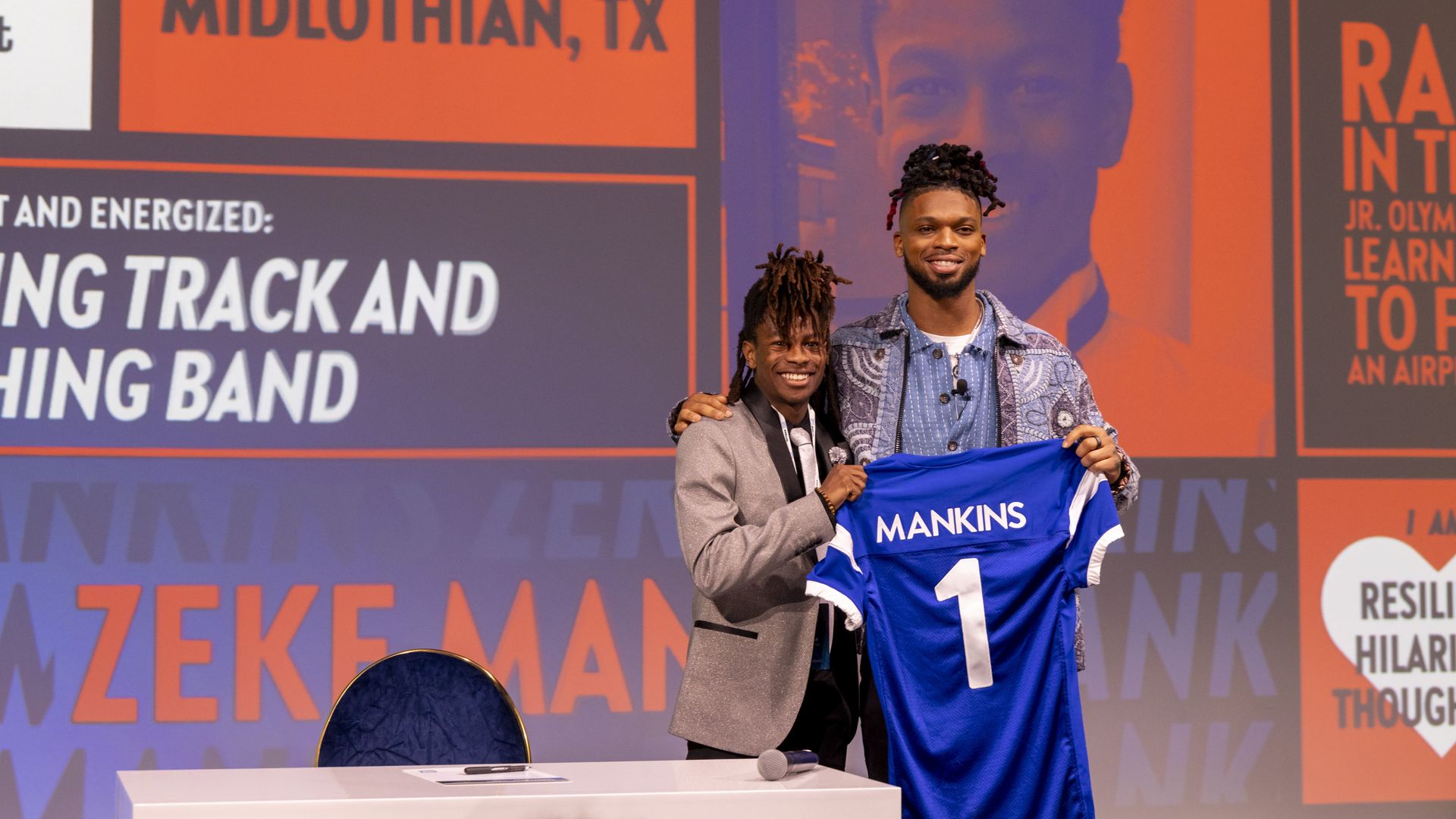 Two men smiling and holding a blue sports jersey with "MANKINS 1" on it, standing in front of a colorful backdrop.