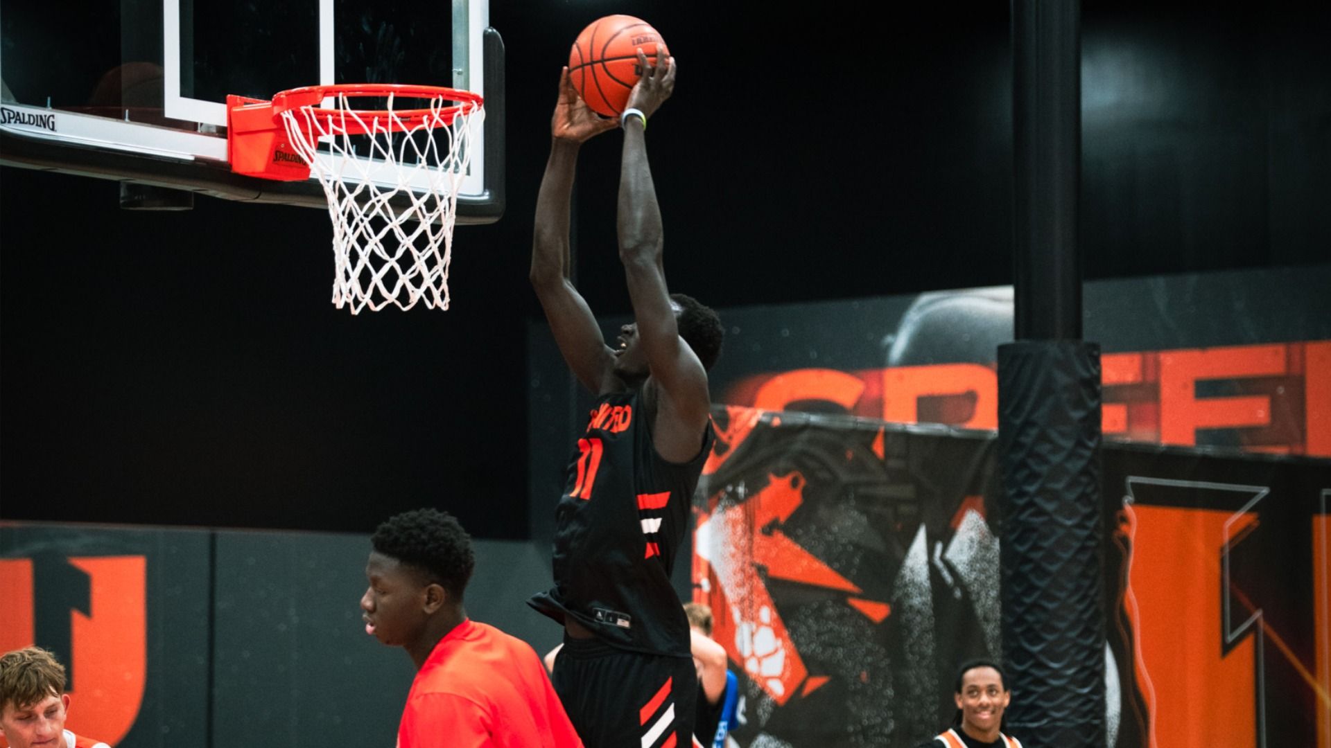A basketball player in a black and orange uniform jumps to dunk a basketball near a red and white hoop in an indoor court with black and orange walls. Other players watch nearby.