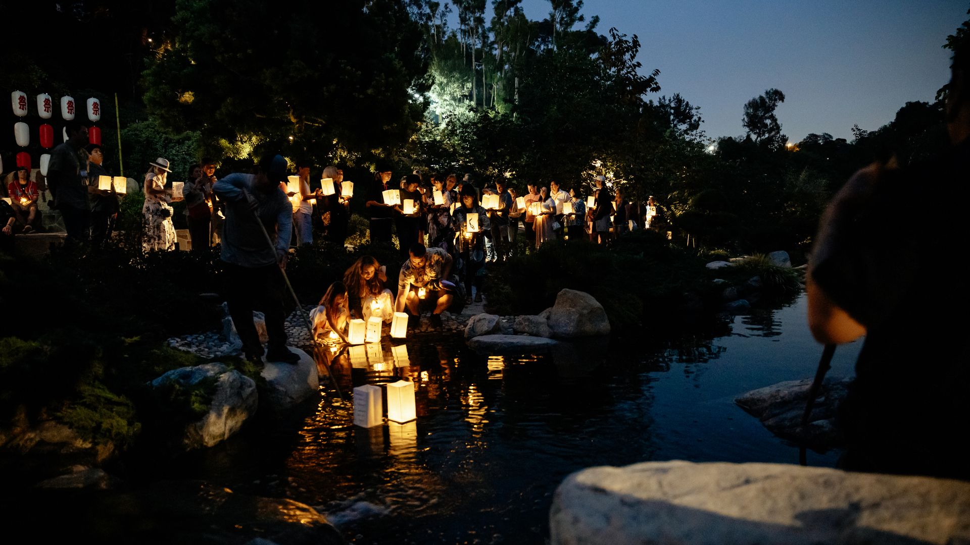 A crowd of people walks along a rocky garden pond at dusk, holding glowing paper lanterns that illuminate faces as lanterns line the water and a row of white and red lanterns hang to the left.