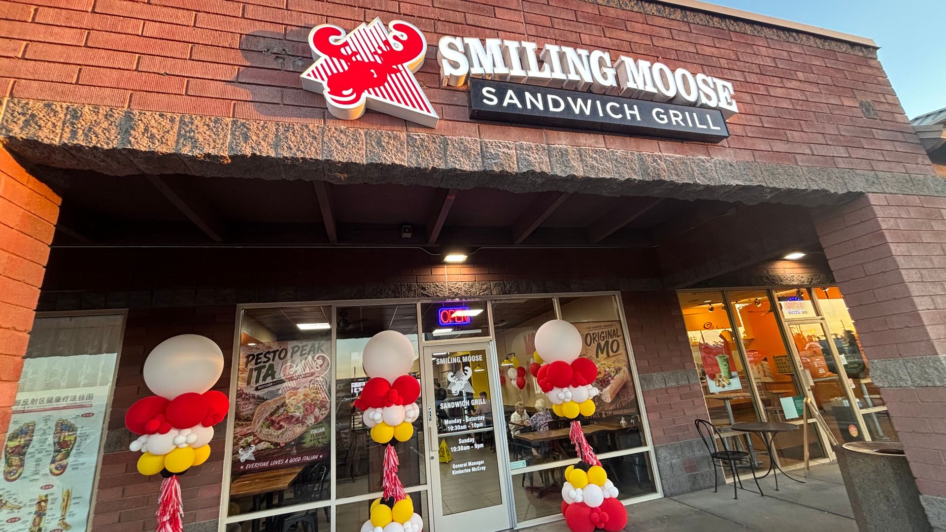 Brick storefront with a large sign reading "Smiling Moose" and "SANDWICH GRILL." Red, white, and yellow balloon column decorations flank the entrance; glass doors show posters under a clear blue sky.