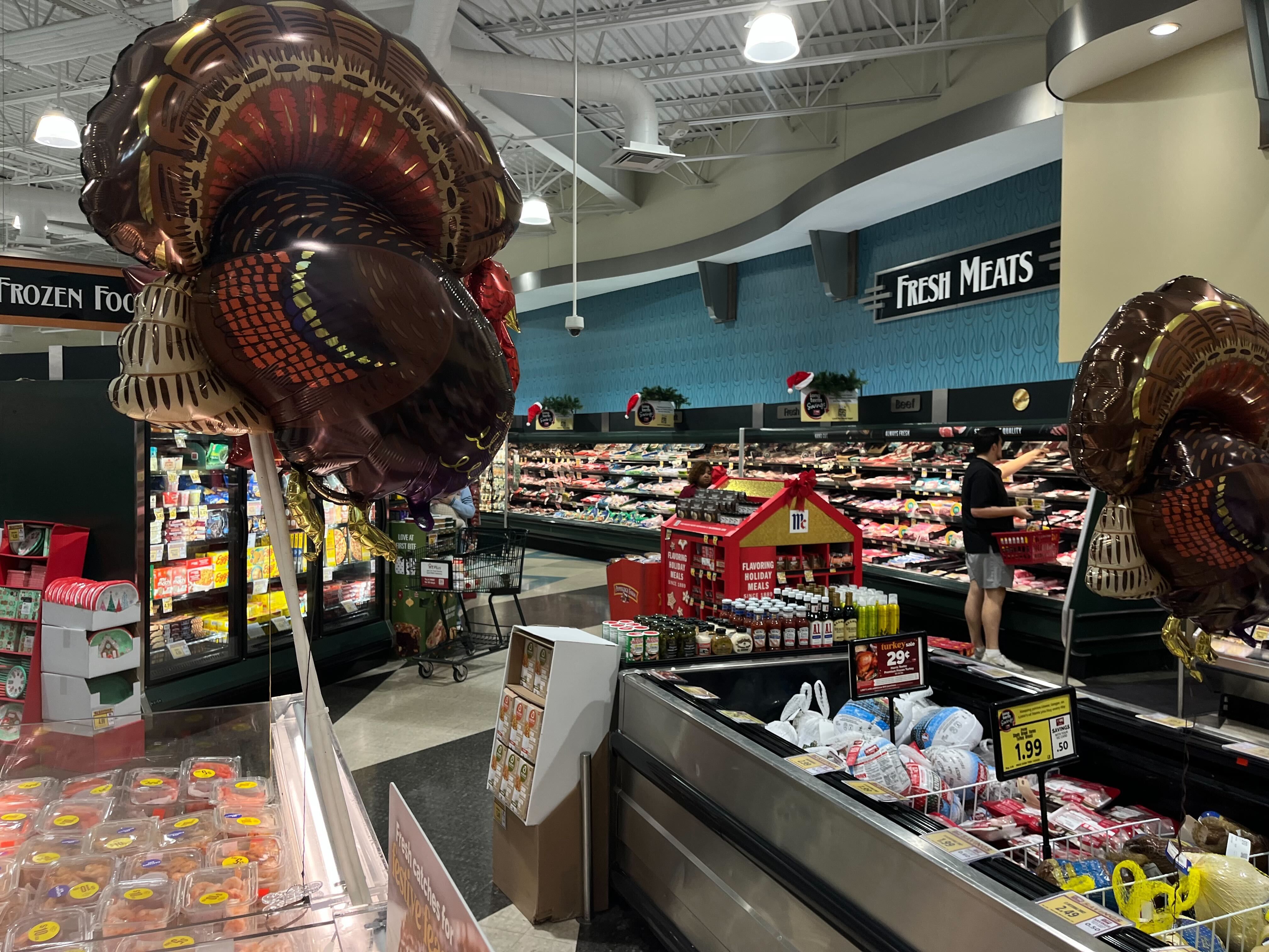 Grocery store meat section with turkey-shaped balloons, turkeys on sale, and holiday meal seasonings on display. Signs say Fresh Meats and Frozen Foods.
