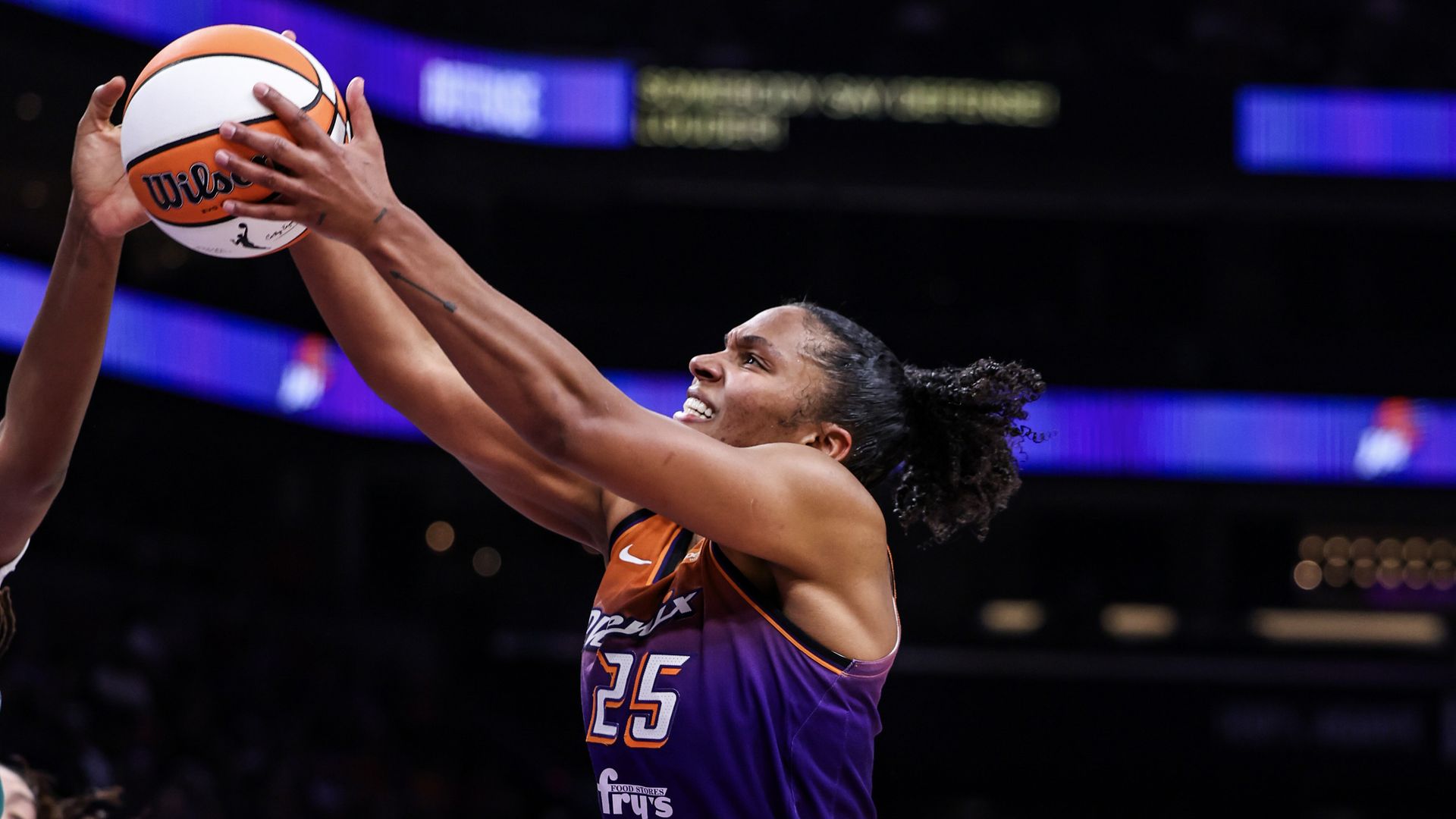 Female basketball player in purple and orange Phoenix jersey (#25) intensely reaching for a Wilson basketball in a game, with blurred crowd and arena lights in the background.