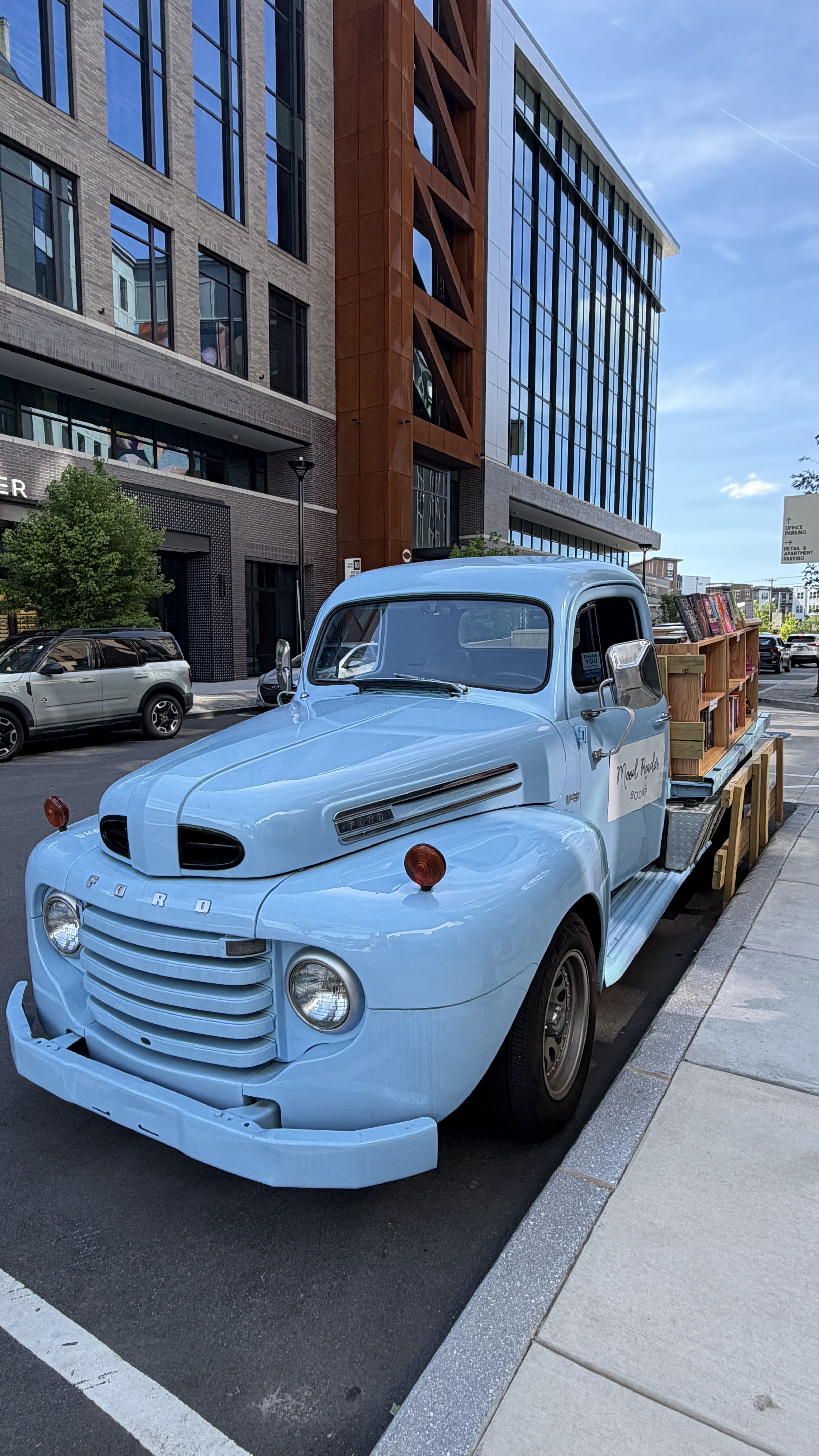 A light blue vintage Ford truck on a city street, its flatbed loaded with wooden crates and books. Modern brick-and-glass buildings tower behind under a clear blue sky.