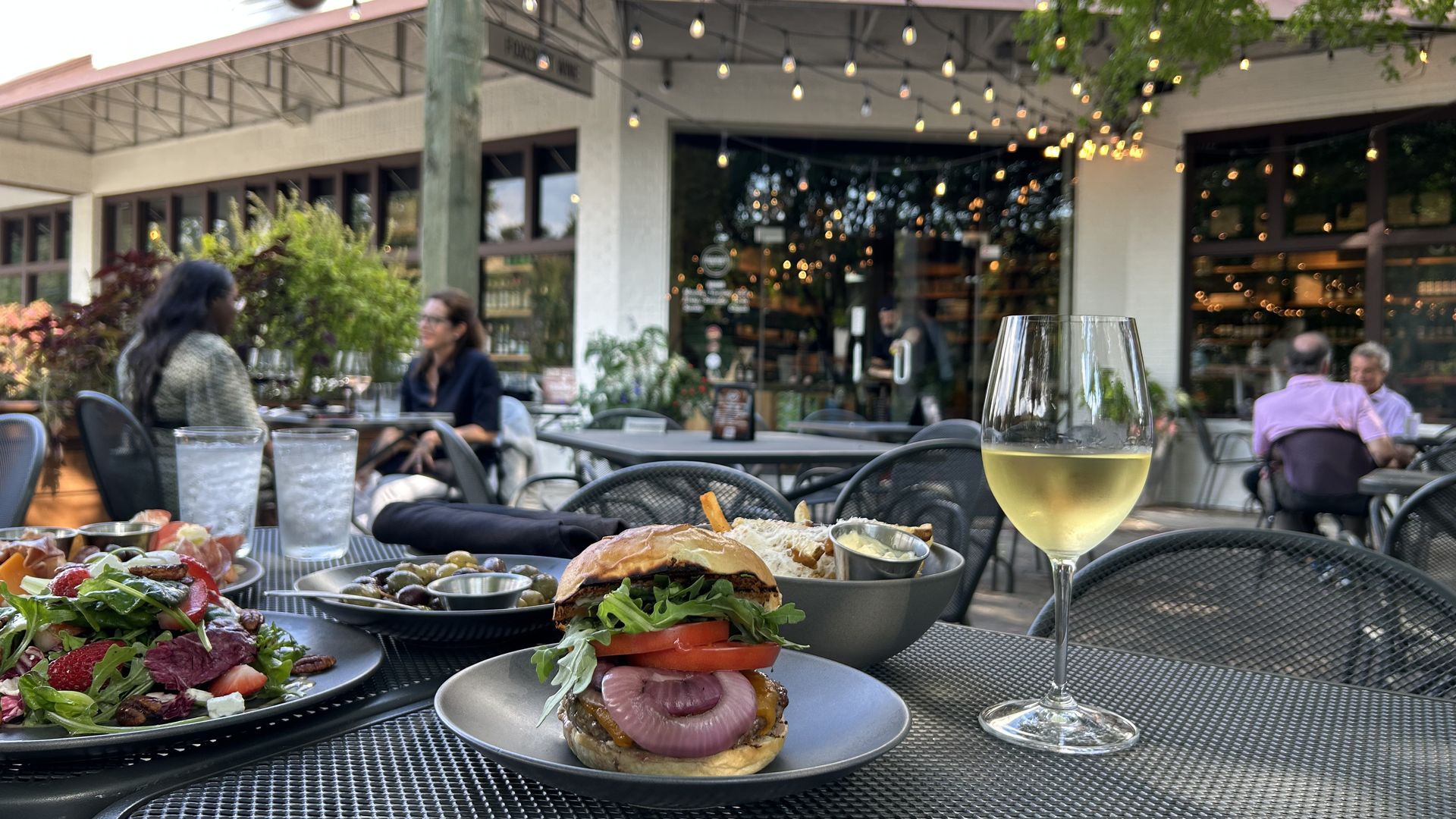Outdoor patio dining at a cafe with string lights, a burger and fries on a plate, salad with strawberries, olives, two glasses of water, and a glass of white wine on a metal mesh table.