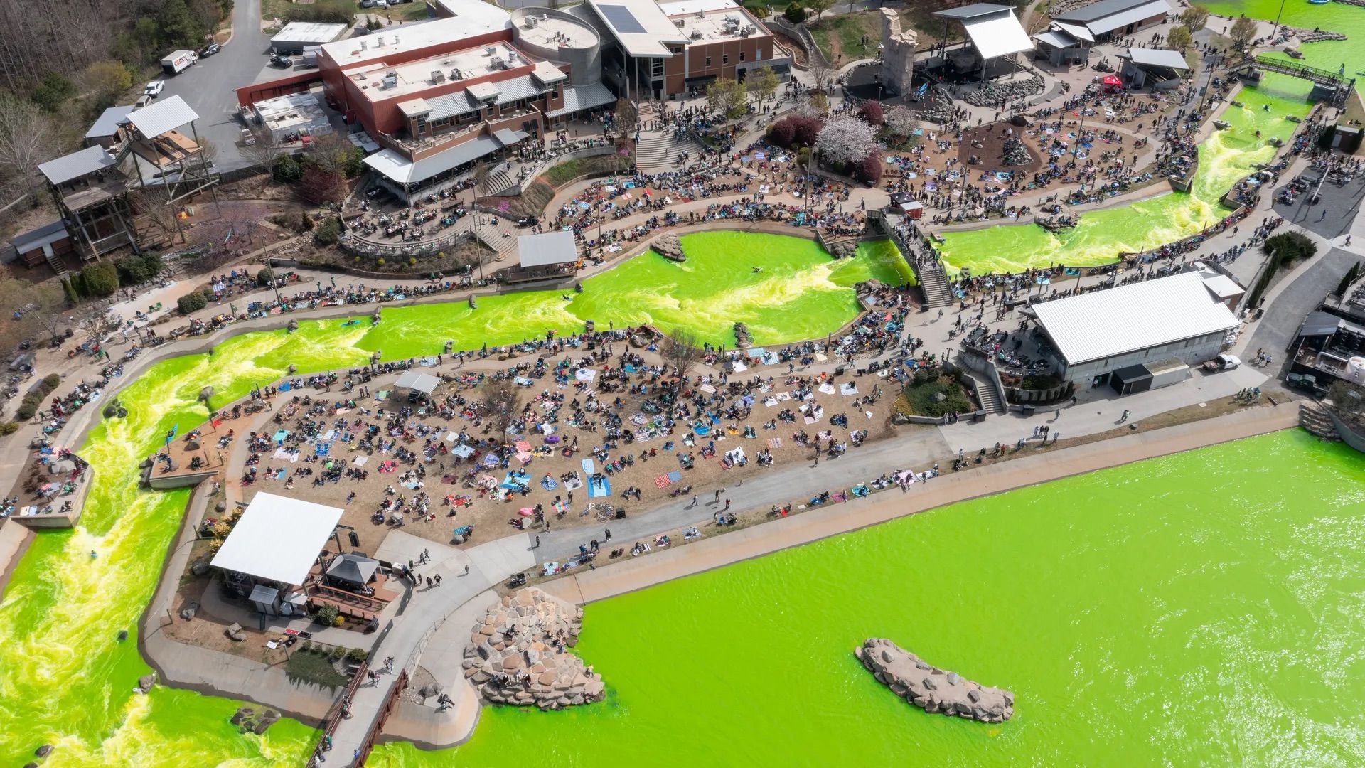 Aerial view of a crowded outdoor event along a neon green man-made river. Bridges, terraces, and white-roofed buildings surround crowds sitting on blankets and strolling the paths.