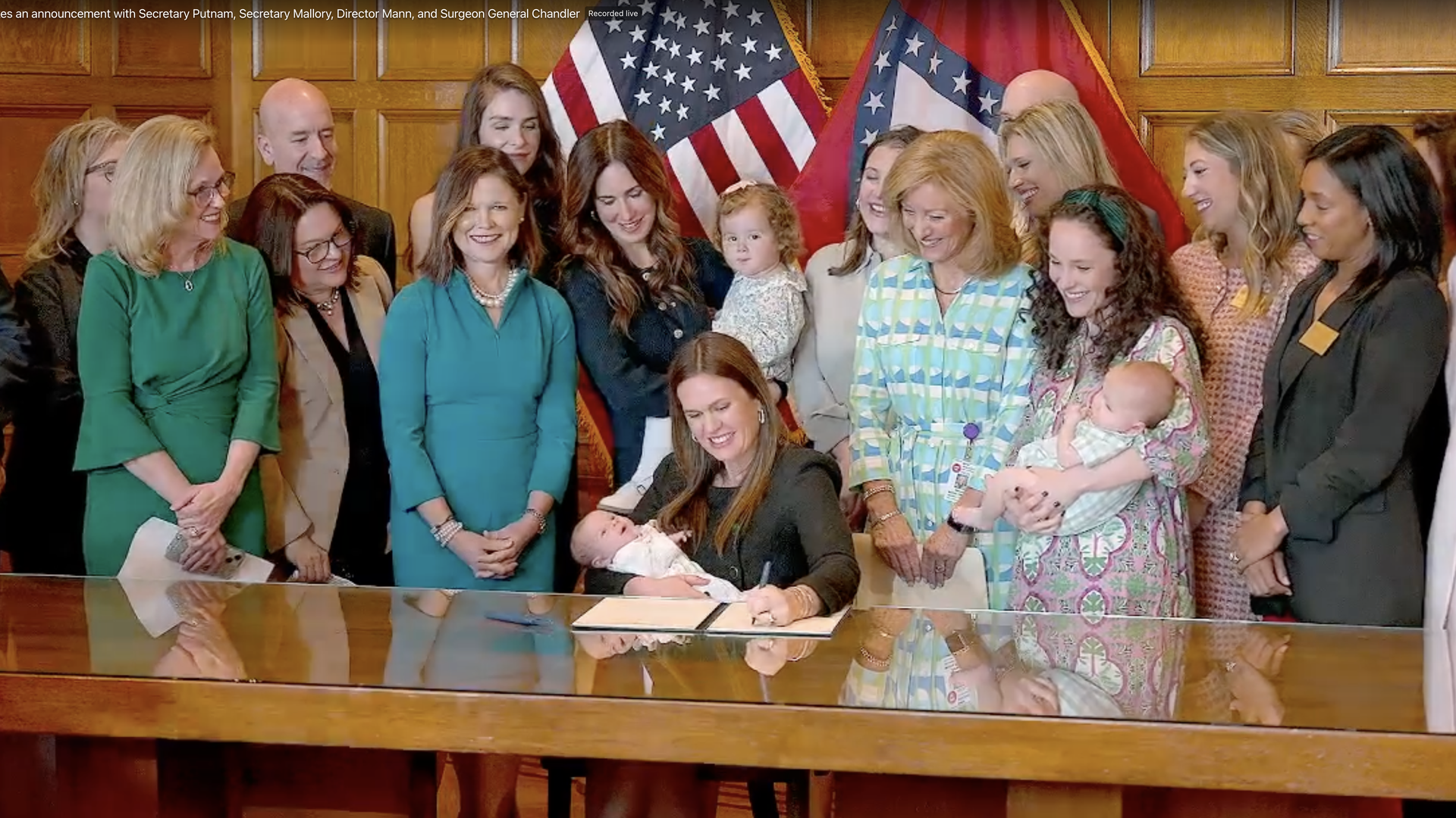 A photo of Gov. Sarah Huckabee Sanders signing an executive order while holding a baby, flanked by women and children.