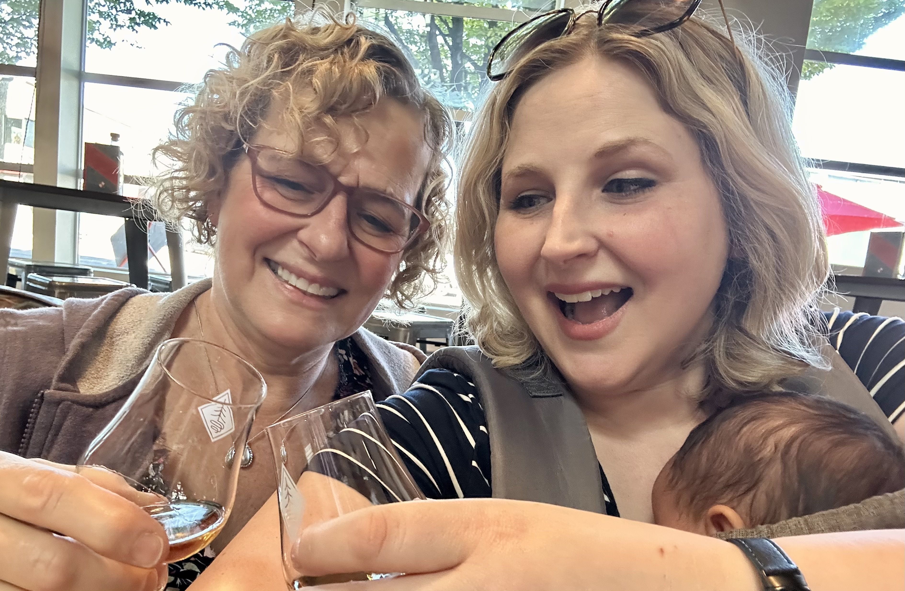 Two women take a selfie in a bright cafe, smiling. Left: curly-haired woman with red glasses; right: blonde with sunglasses on her head. A child and wine glasses are in the foreground.