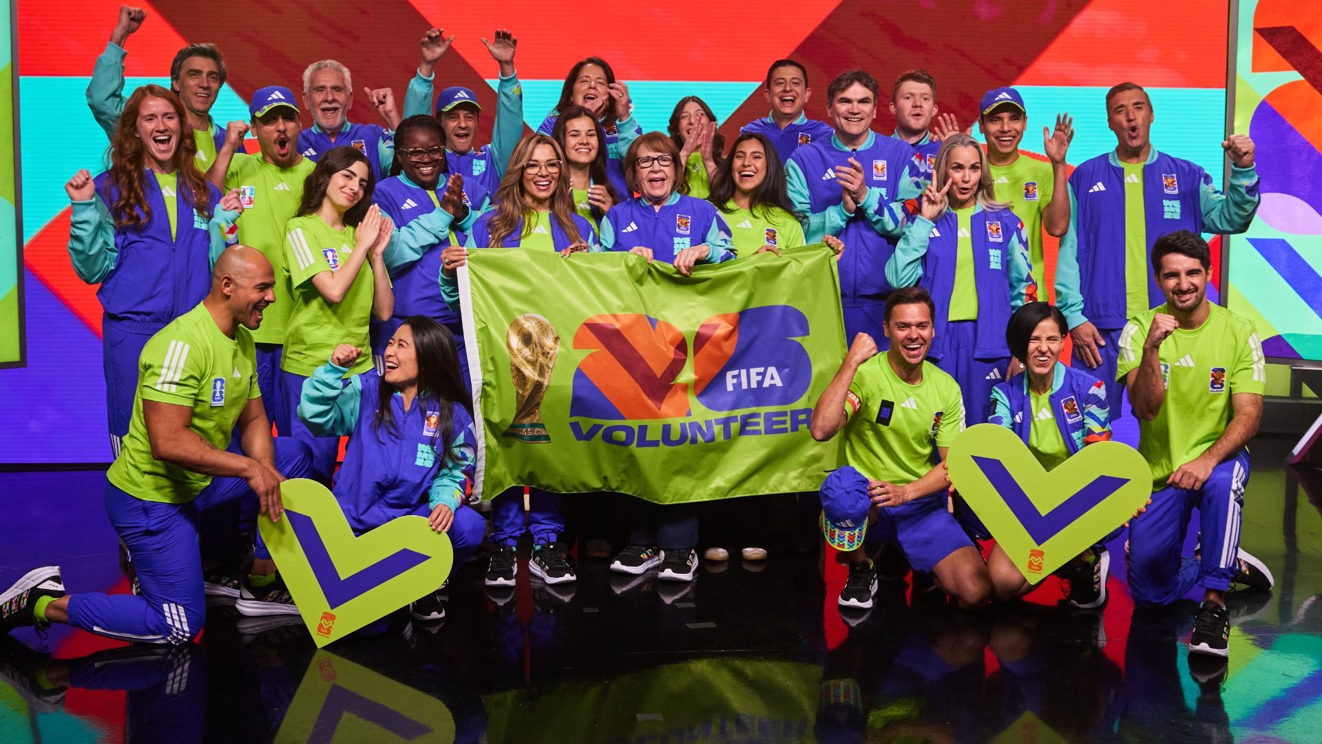 Group of diverse FIFA volunteers in neon lime and blue outfits on a colorful stage, holding a bright green banner with FIFA VOLUNTEER and a trophy image, cheering and raising fists.