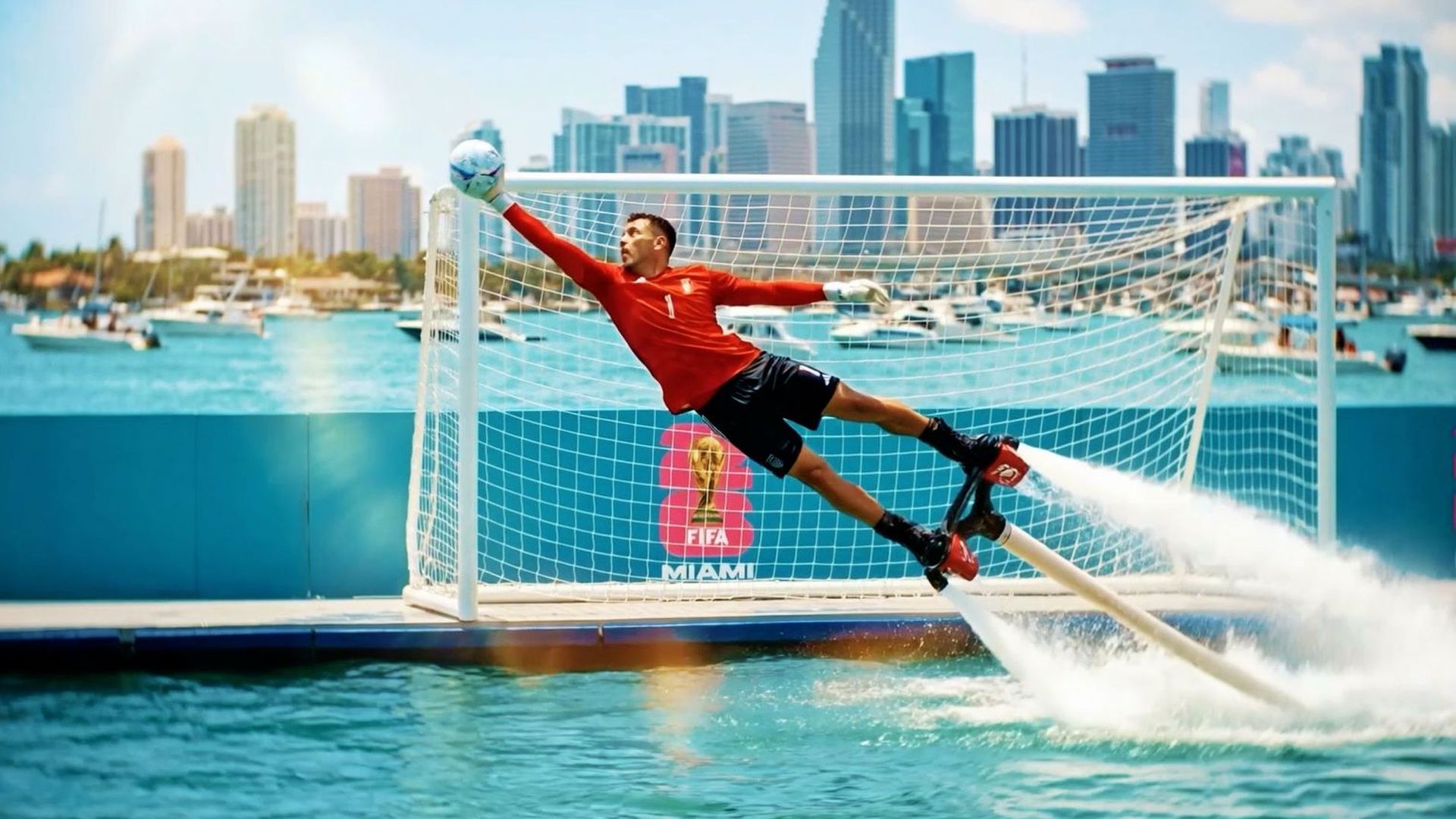 Red-jersey goalkeeper dives horizontally to save a ball at a water-soccer goal; water jets spray from his feet as a city skyline, boats, and a blue Miami barrier backdrop.