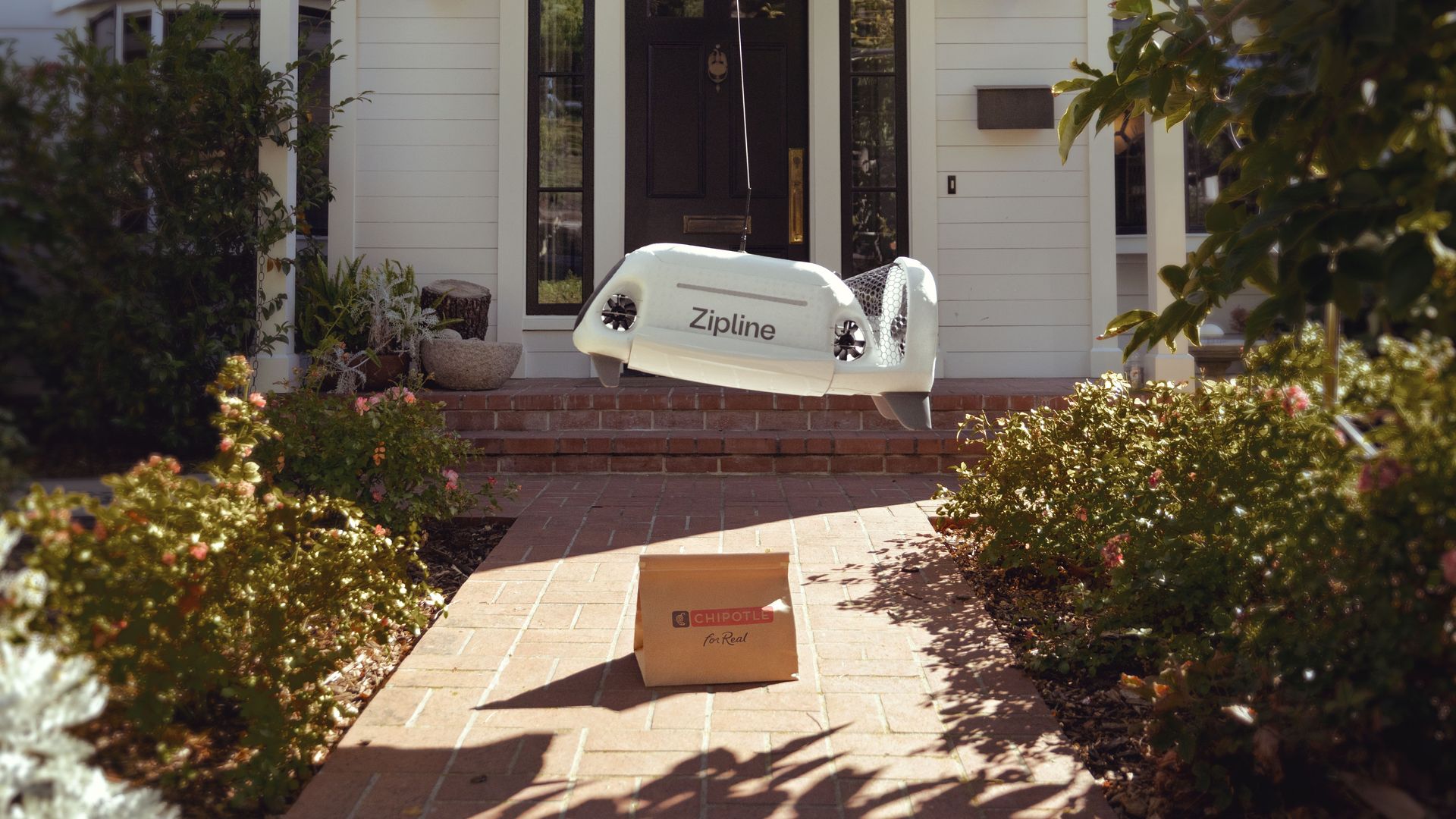 A white Zipline delivery drone suspended by a tether leaves a brown bag of Chipotle food on the brick path in front of a home.