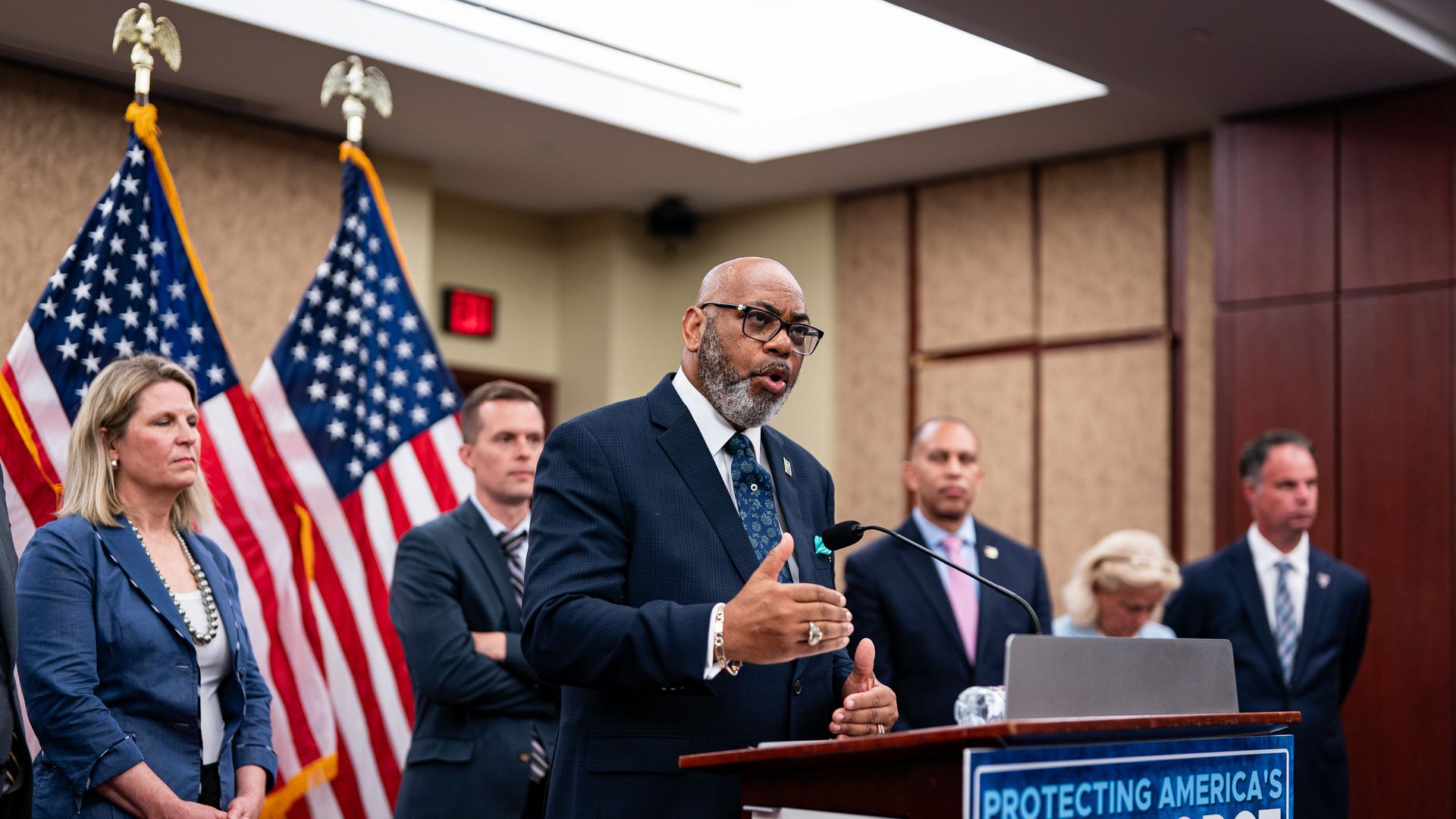 Everett Kelley wearing a blue suit and tie with a white shirt, speaking in front of several people and American flags. 