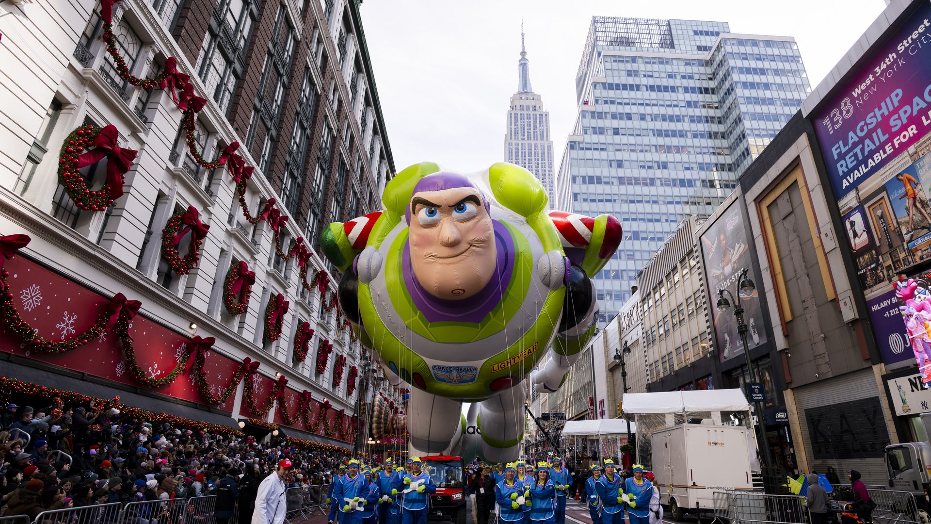 A large balloon of the Buzz Lightyear character, featuring fair skin, blue eyes, and an outline of a swirl on his chin, which gives him a cleft chin, with the Empire State Building and Macy's department store in the background during the 2025 Macy's Thanksgiving Day Parade on November 27 in NYC.