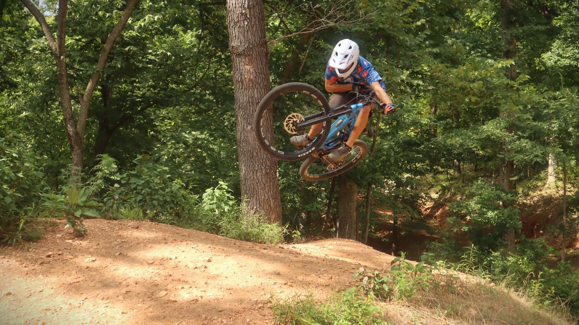 A person on a mountain bike gets a lot of air in a jump over a dirt hill with woods in the background.