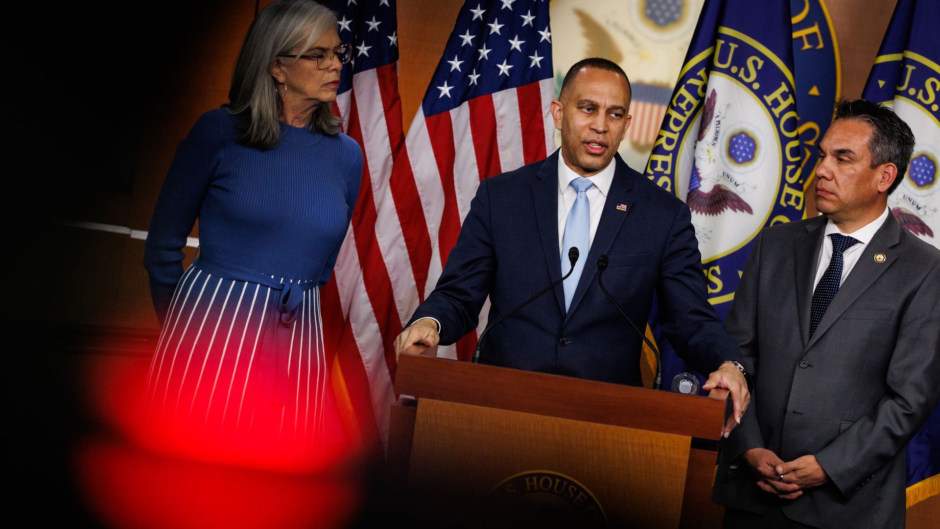 Three people stand at a podium with U.S. flags and congressional seals behind them. Left: a woman in a blue top and striped skirt. Center: a man in a navy suit speaking. Right: a man in a gray suit.