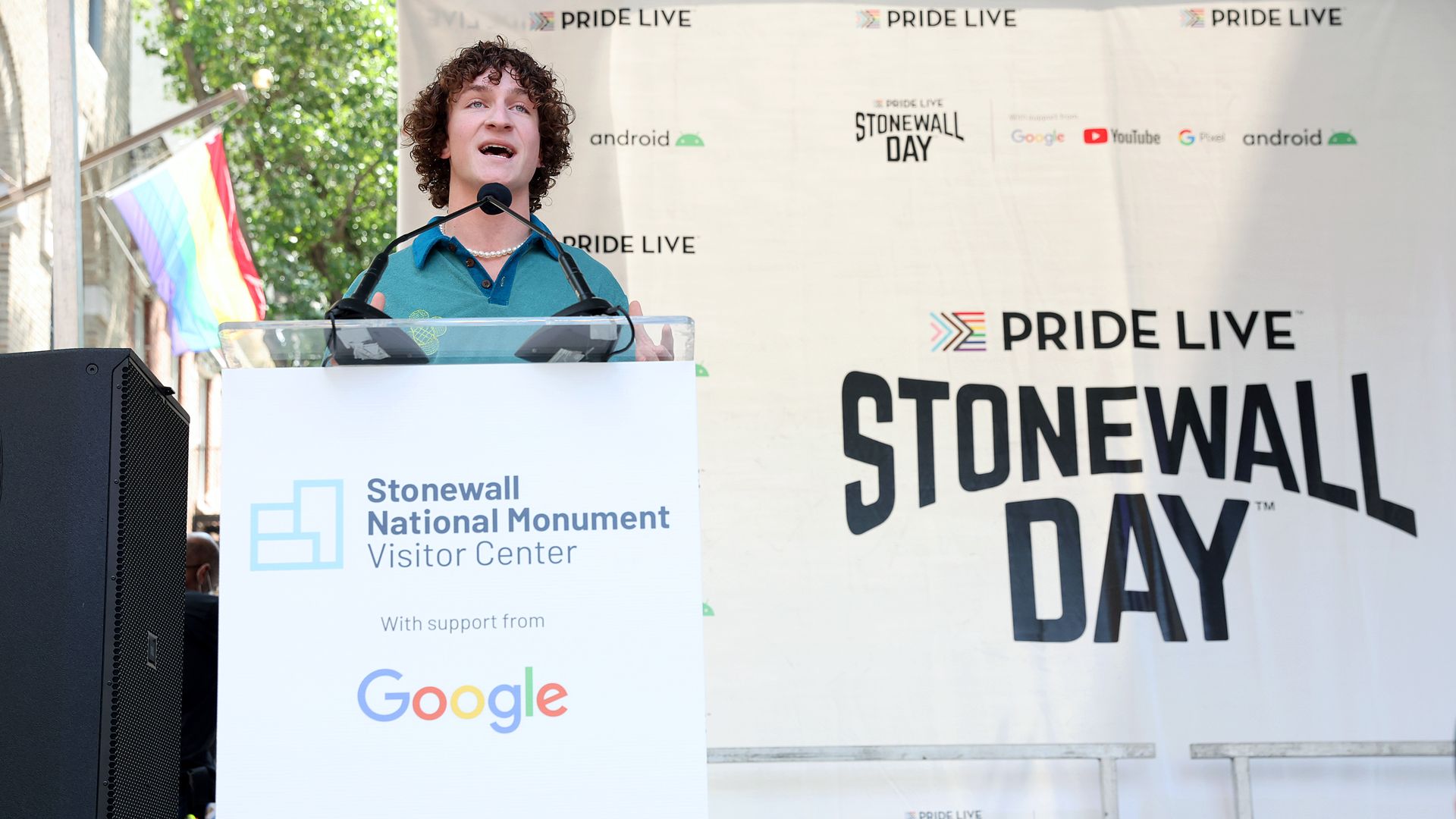 A person stands at a lectern that reads "Stonewall National Monument Visitor Center with support from Google" with a pride flag and sign that reads "Pride Live Stonewall Day" in the background.