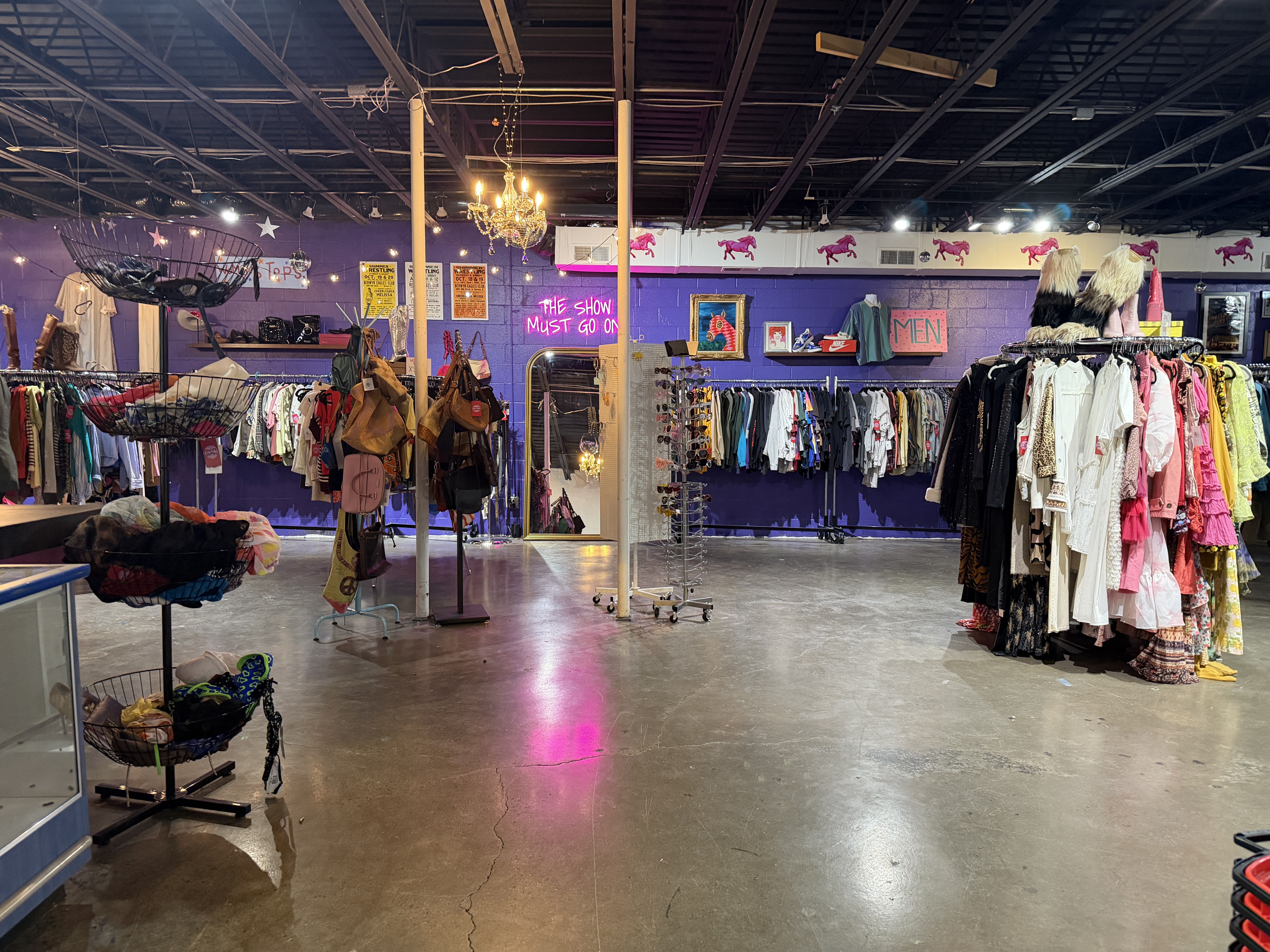 Interior of a vintage thrift store: purple walls, racks full of colorful dresses and jackets, bags on stands, a chandelier, neon pink sign says "THE SHOW MUST GO ON", and a "MEN" section in back.