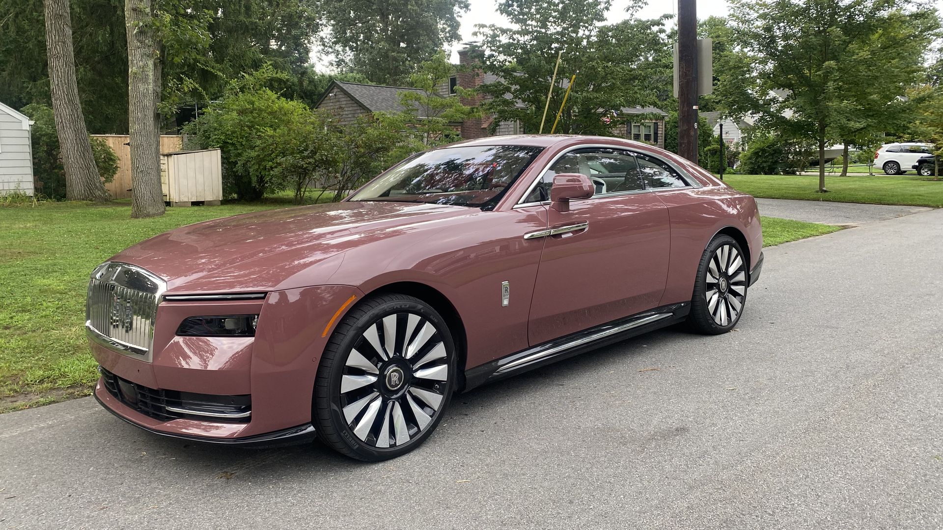 A photo of a reddish/pinkish two-door Rolls-Royce Spectre electric coupe, parked on a suburban street.