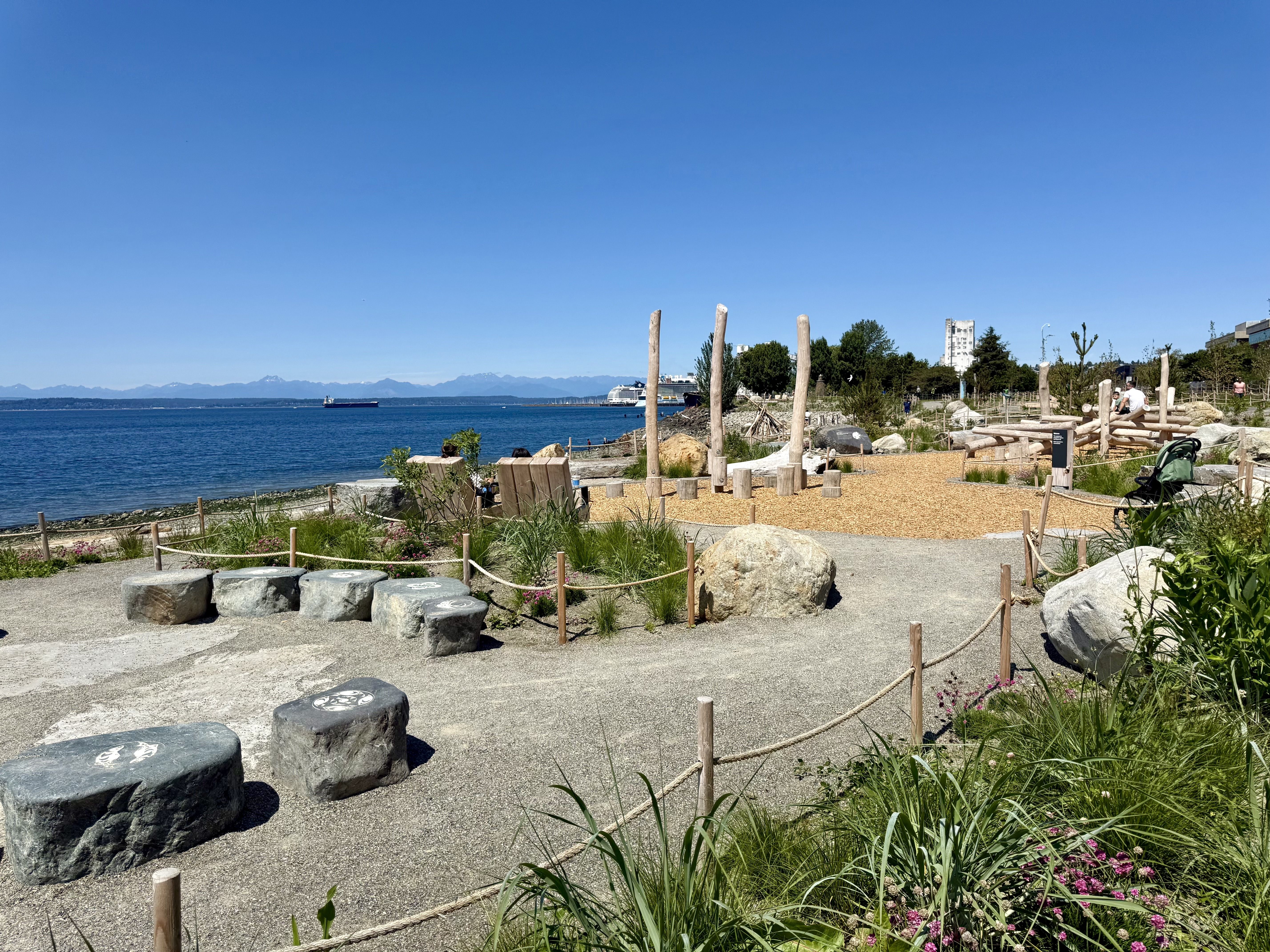 Sunny seaside park with gravel paths, wooden posts, rope barriers, and large stone seats. Plants and pink flowers frame the scene as calm water, a ship on the horizon, and buildings appear.