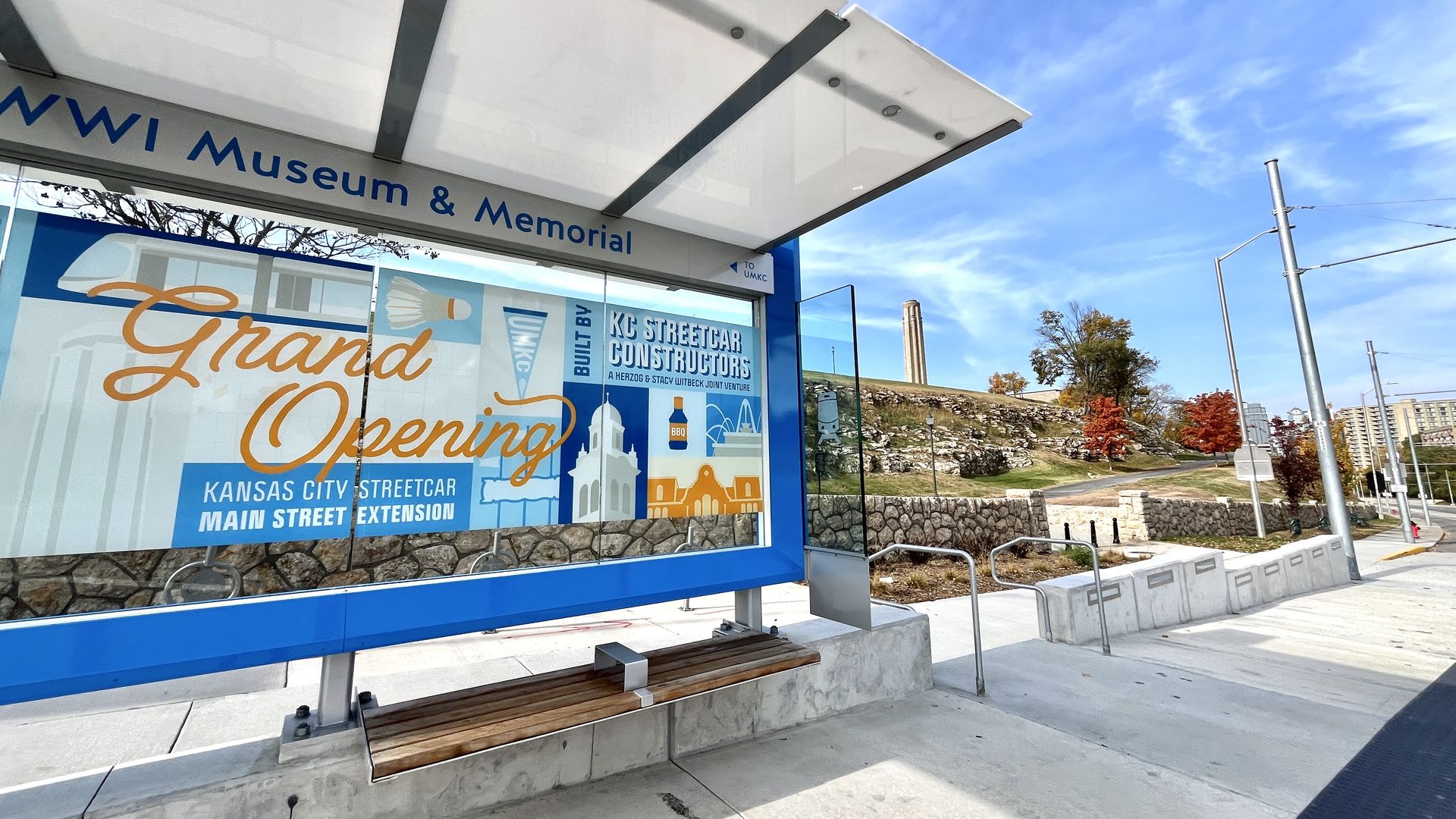 Bus shelter with a colorful sign announcing the grand opening of Kansas City Streetcar Main Street Extension, clear blue sky, trees with autumn leaves, and the Liberty Memorial Tower in the background.