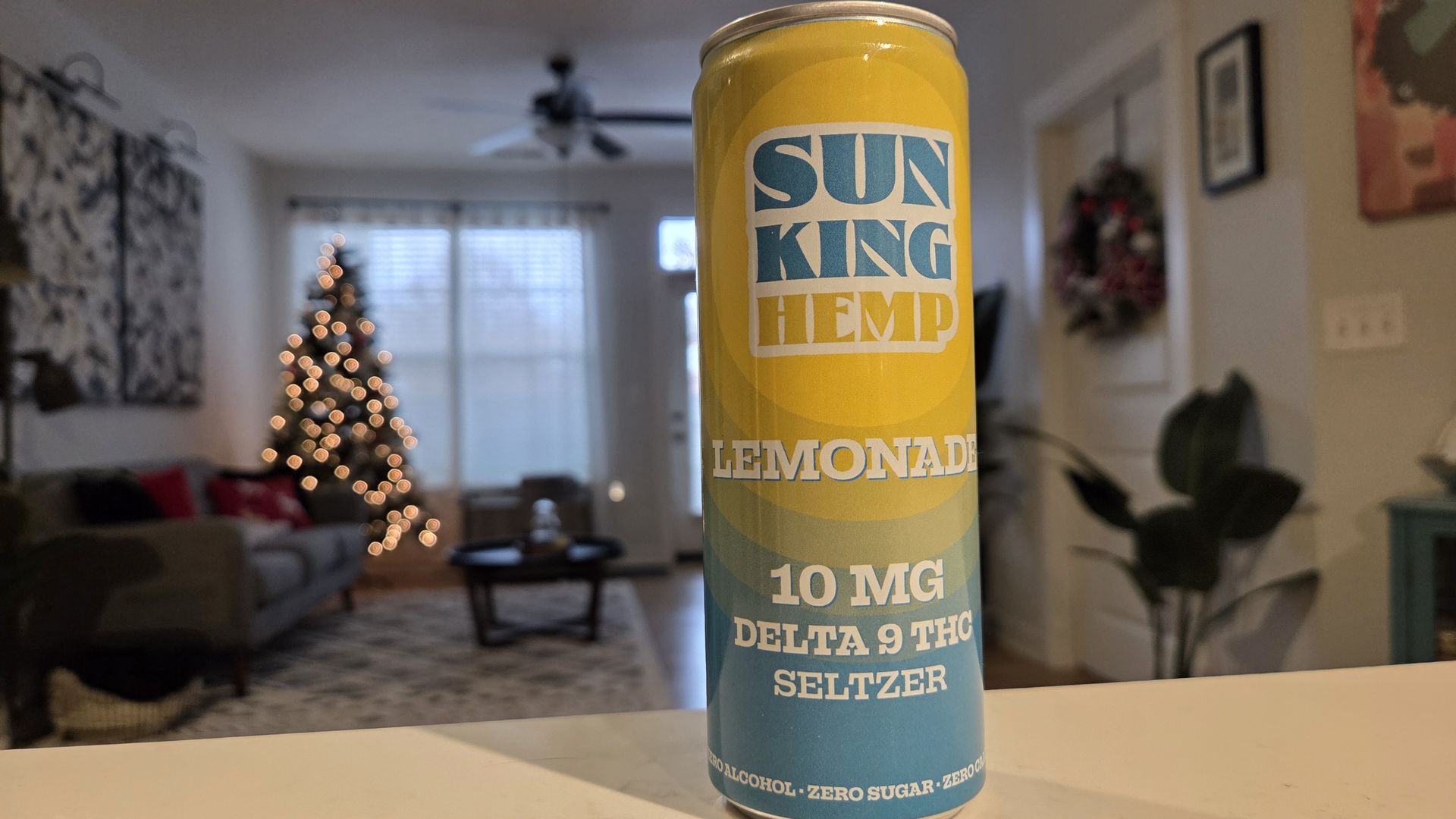 A yellow and blue can of Sun King Hemp Lemonade with 10 mg Delta 9 THC seltzer, placed on a white surface in a cozy living room with a Christmas tree and decorations in the blurred background.