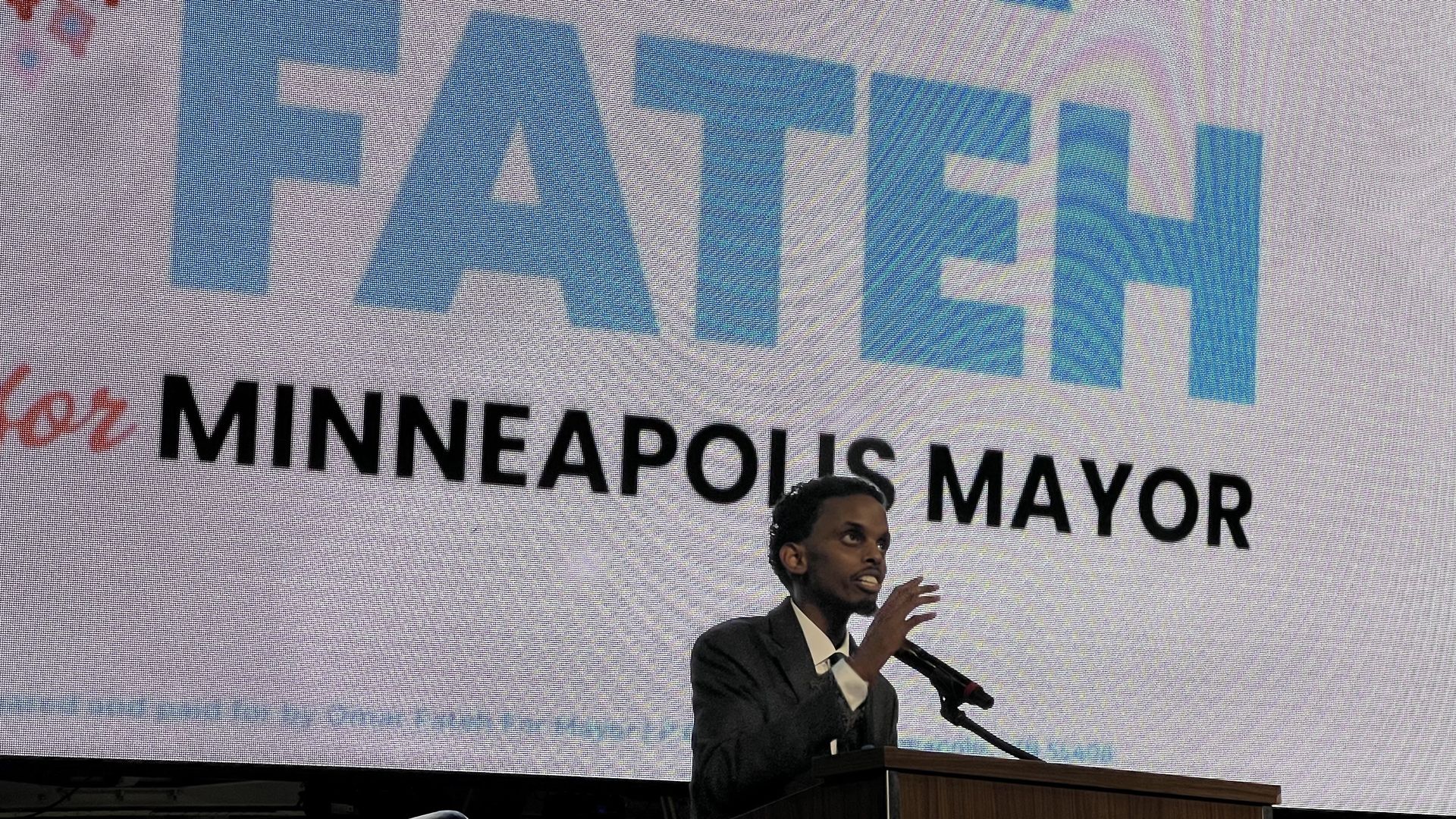 A man in a dark suit speaking at a podium with a microphone in front of a large screen displaying the text "FATEH for MINNEAPOLIS MAYOR" in blue and black letters.