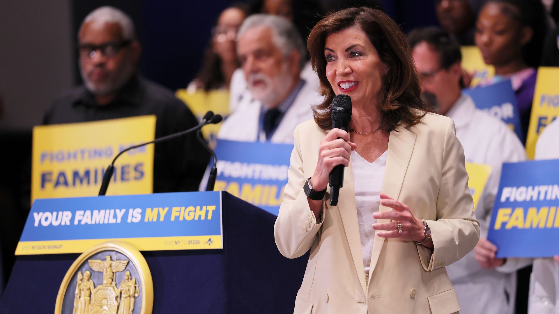 New York Gov. Kathy Hochul wears a tan blazer and holds a microphone in front of a podium with a sign that reads "Your family is my fight." 