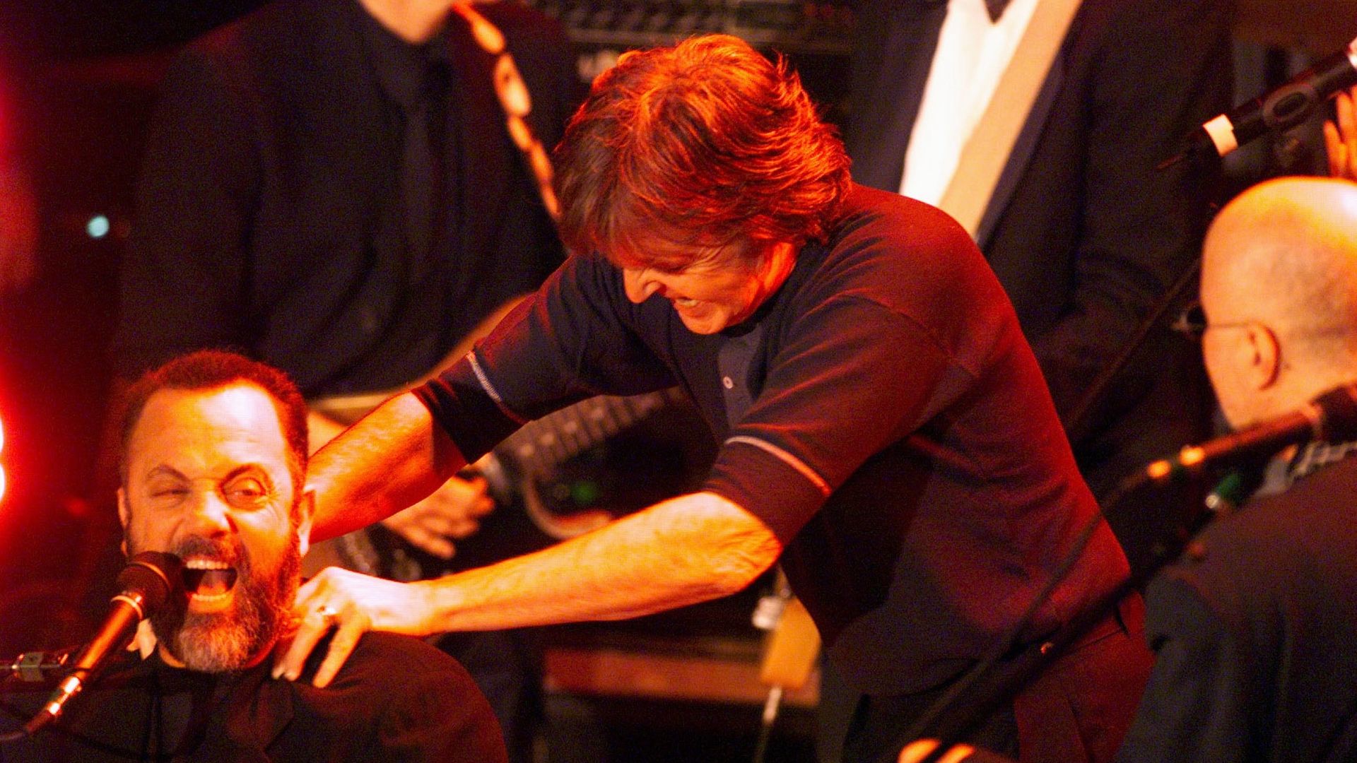 Paul McCartney playfully grabs Billy Joel by the shoulders from behind as Joel performs at the piano on stage.