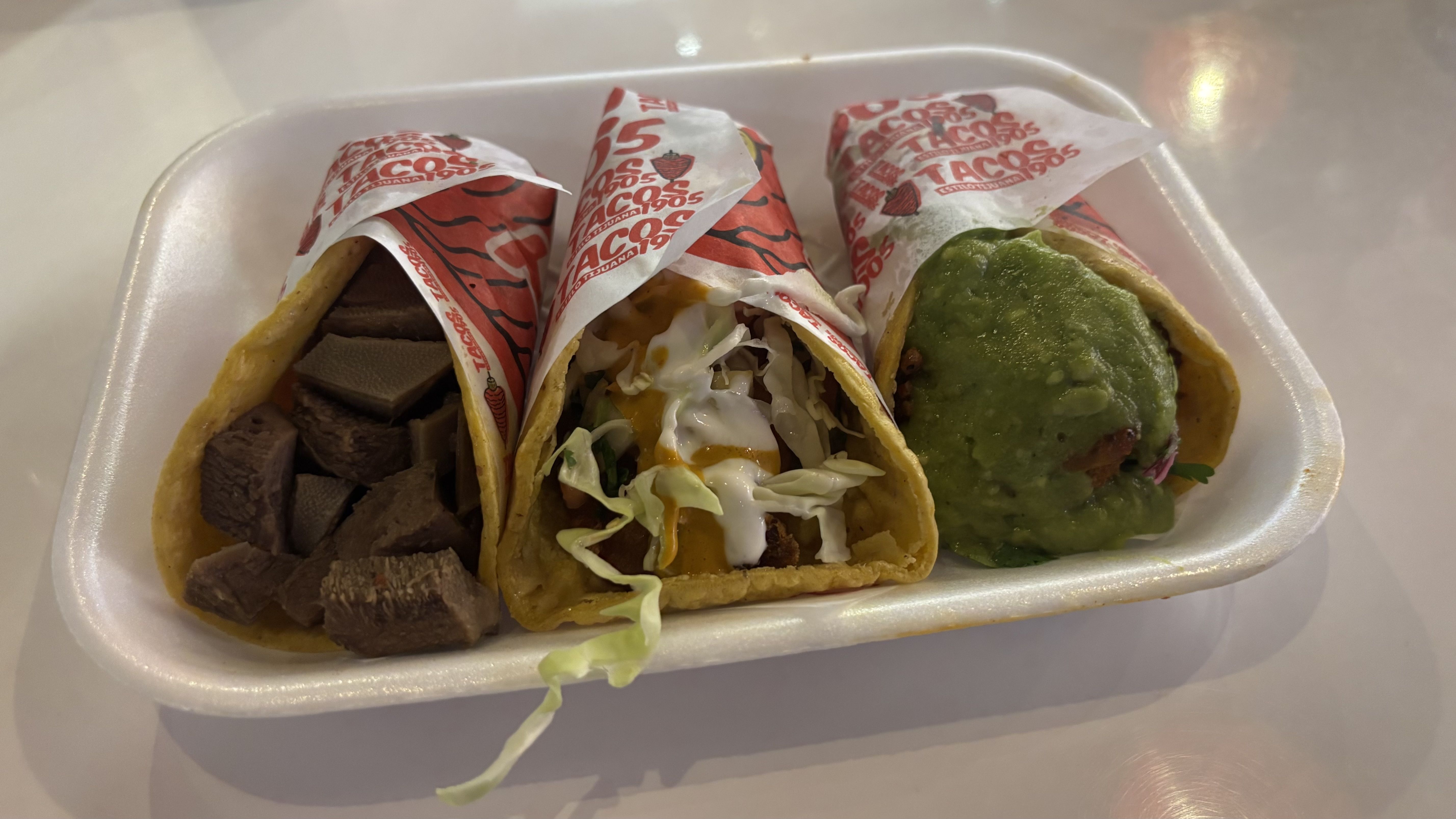 Three tacos in a white foam tray with red and white wrappers. Left taco with diced meat, middle with shredded cabbage and sauces, right topped with green guacamole.