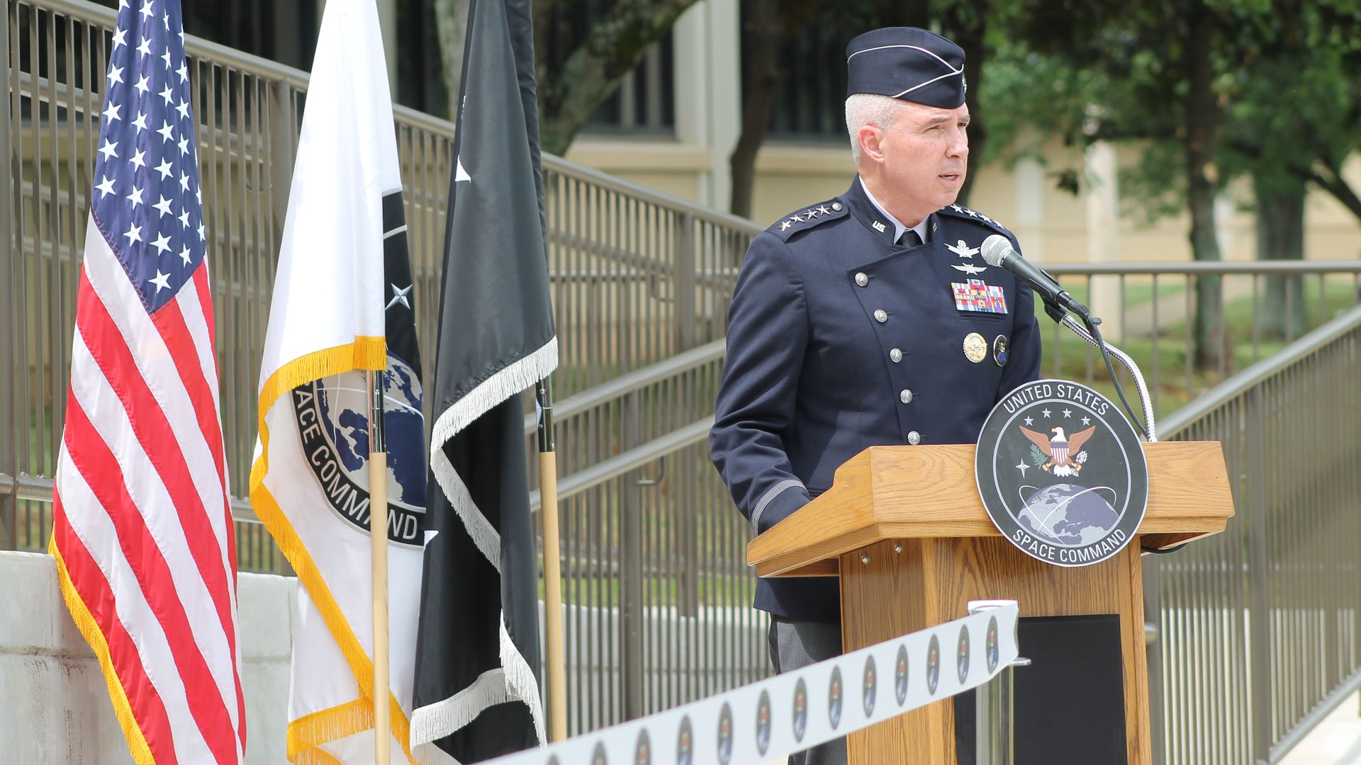 Air Force officer in dark blue dress uniform speaks at a podium with the Space Command seal, flanked by American and other flags outdoors, with green trees in the background.