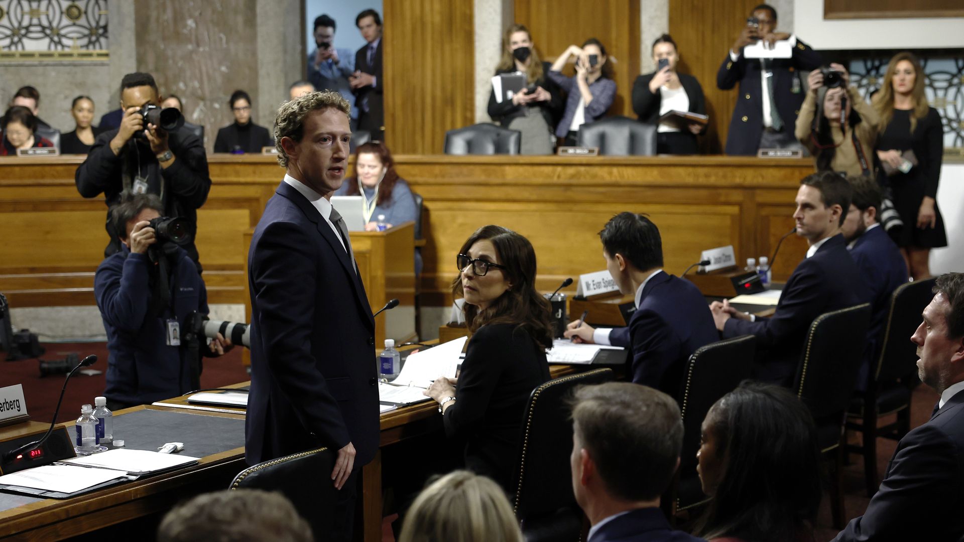 A photo of a Congressional hearing where Mark Zuckerberg stands and turns behind him to speak. Other CEOs are seen in the photo as well as photographers. 