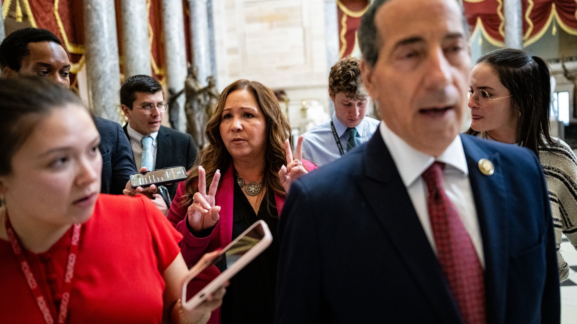 Rep-elect Adelita Grijalva, wearing a pink blazer, walks with reporters, aides and a fellow member in a large white marble chamber with gray marble columns and red curtains.