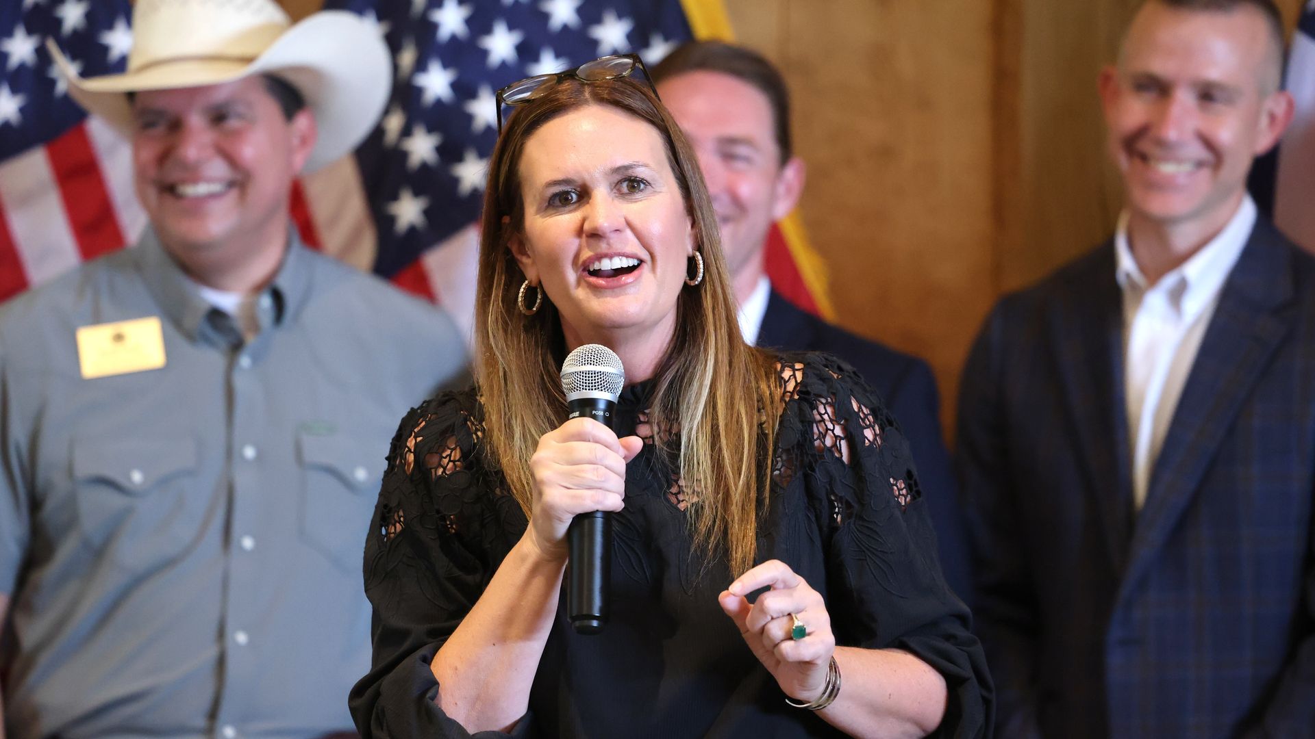 A smiling woman in a black lace top speaks into a microphone, with glasses perched on her head. Behind her, American flags and smiling team members suggest a political or civic event.