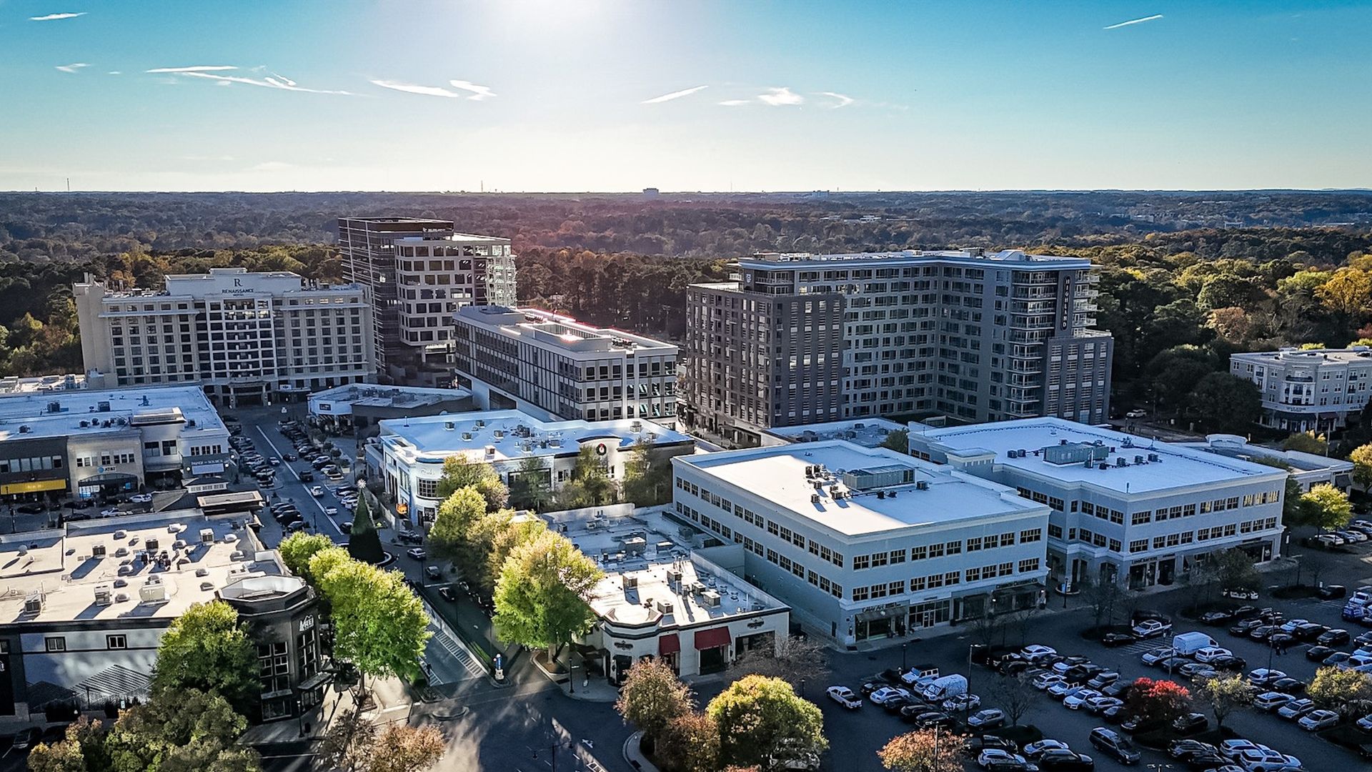Aerial view of a modern city center with white rooftops, high-rise buildings, and tree-lined streets, extending to a dense forest under a clear blue sky.