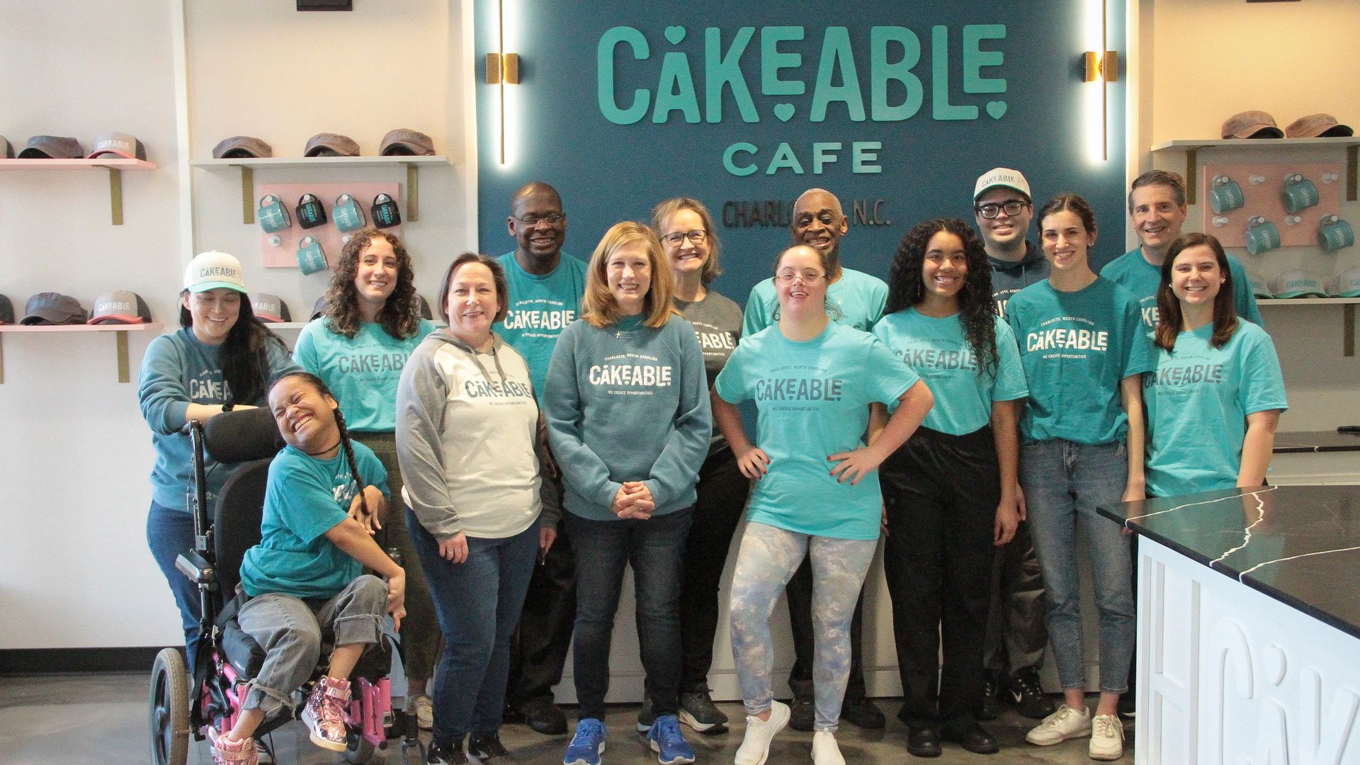 group of 14 people stand in front of a sign tha treads cakeable cafe