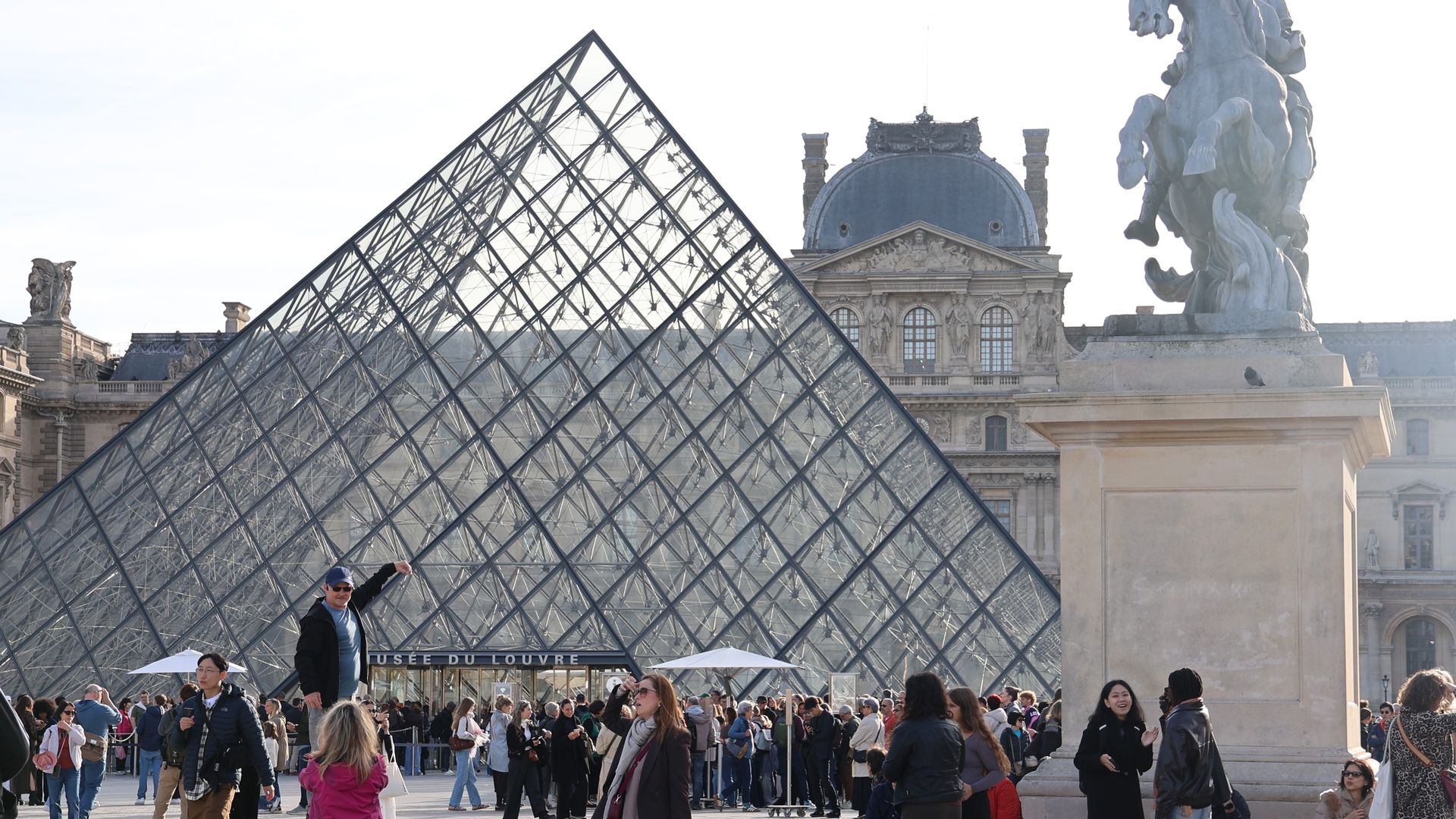 Crowds of people stand around the Louvre Pyramid near a statue of a horse.