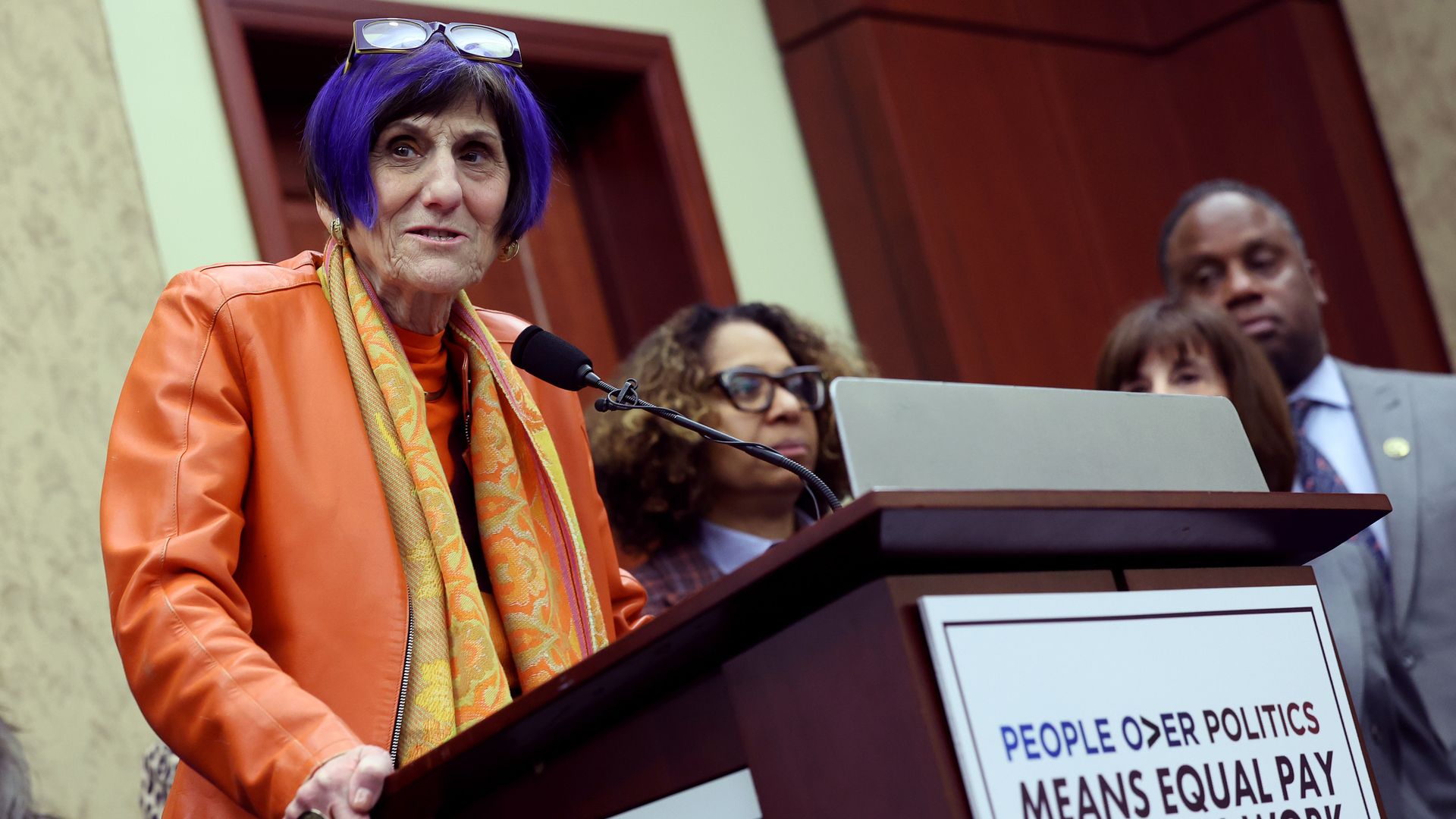 Rep. Rosa DeLauro wears an orange jacket as she stands behind a podium with a placard that says people over politics means equal pay.