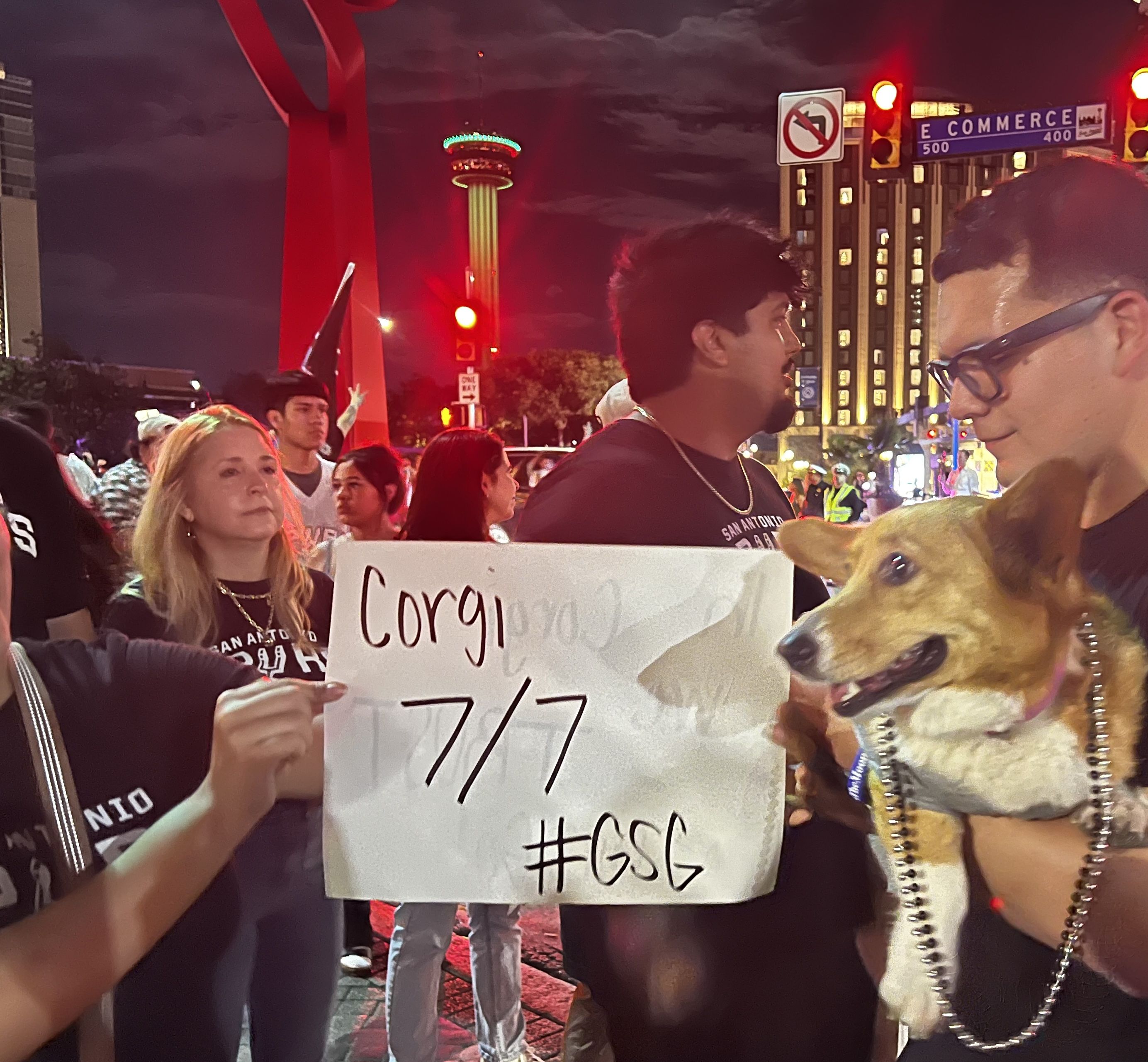 Nighttime street celebration with people holding a white handwritten sign; red lights illuminate faces, a blue E COMMERCE street sign in the background, and a tall tower with a green-lit ring.