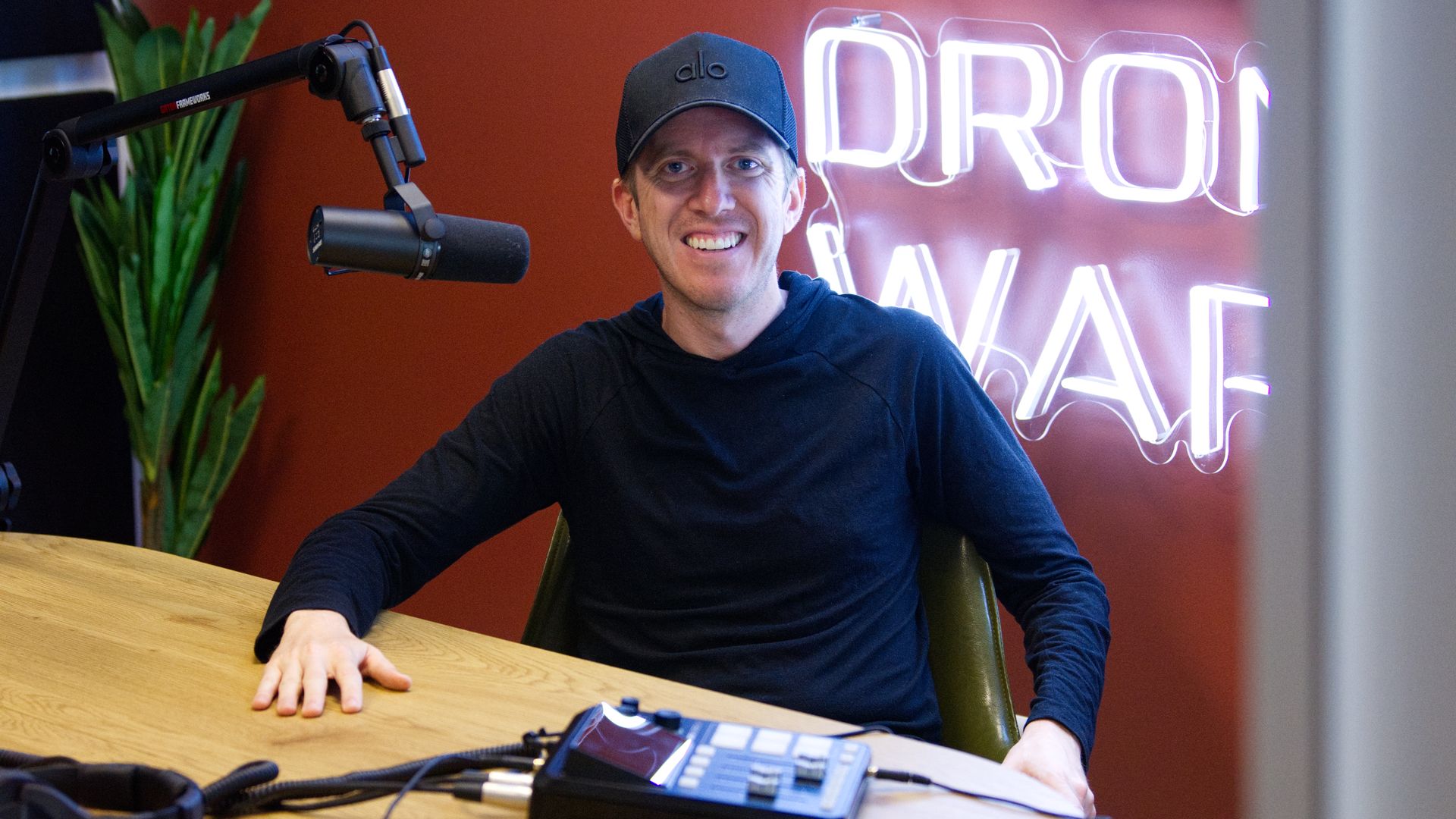 A man in all black, including a hat, sits in a red podcast recording room. A plant is seen to the left; a neon sign to the right.