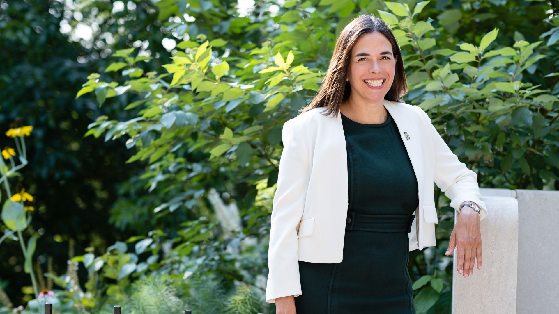 photo of a woman in a white blazer over a black dress in front of a lot of foliage