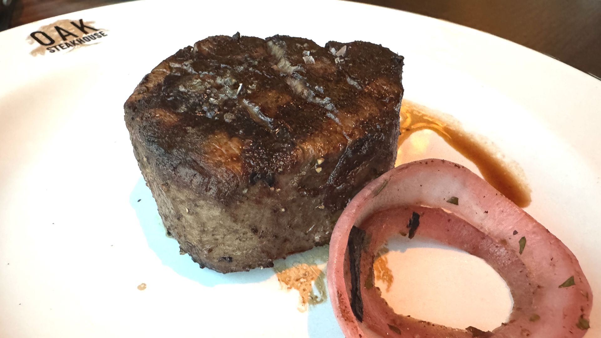 A grilled filet mignon sits on a white plate with the Oak Steakhouse logo, accompanied by a grilled red onion ring and steak juices.
