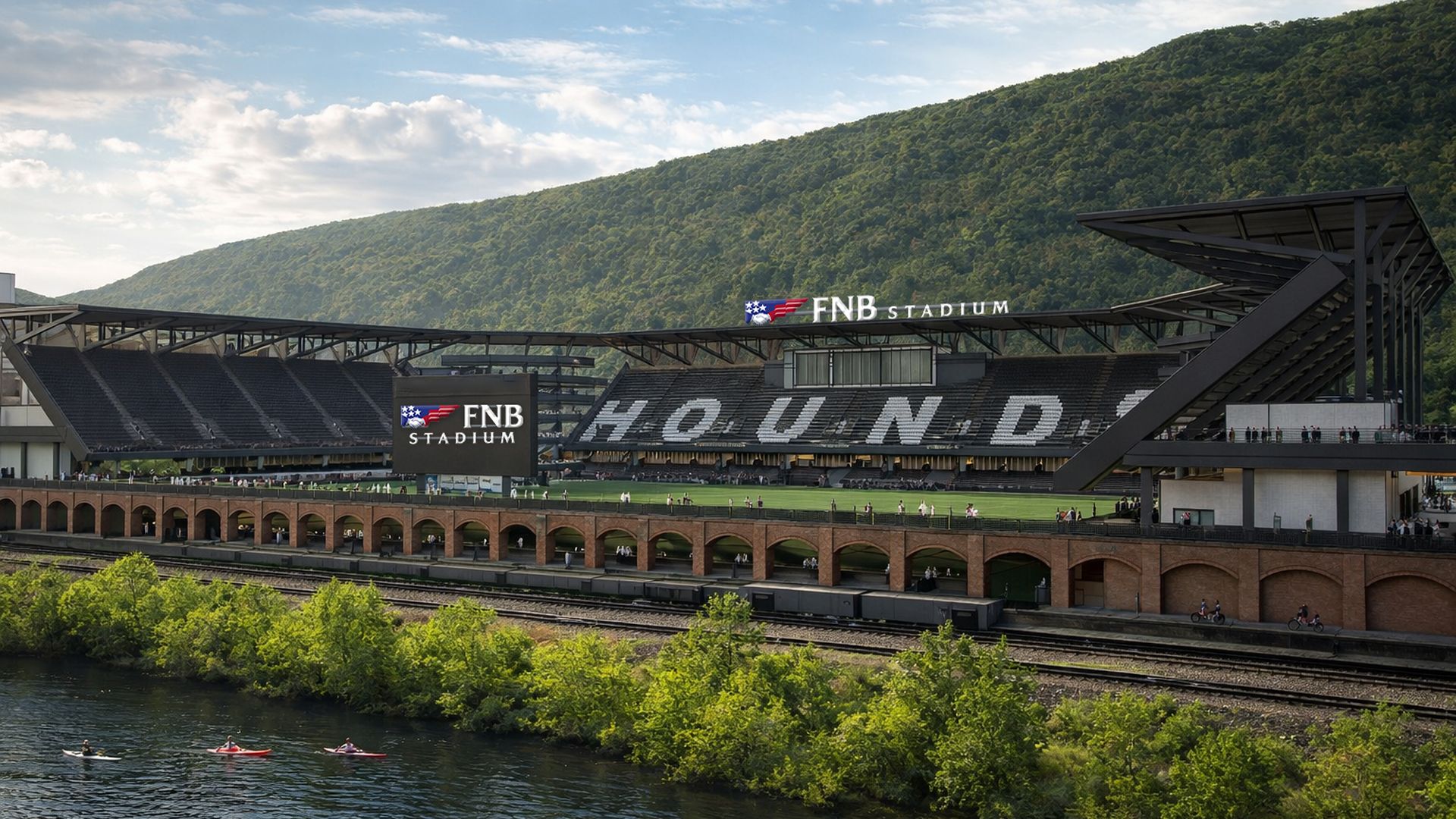 Wide view of FNB Stadium with green hillside and river in front. Bleachers spell "HOUND" across seats; brick arches line the riverbank; kayakers and people on the field.