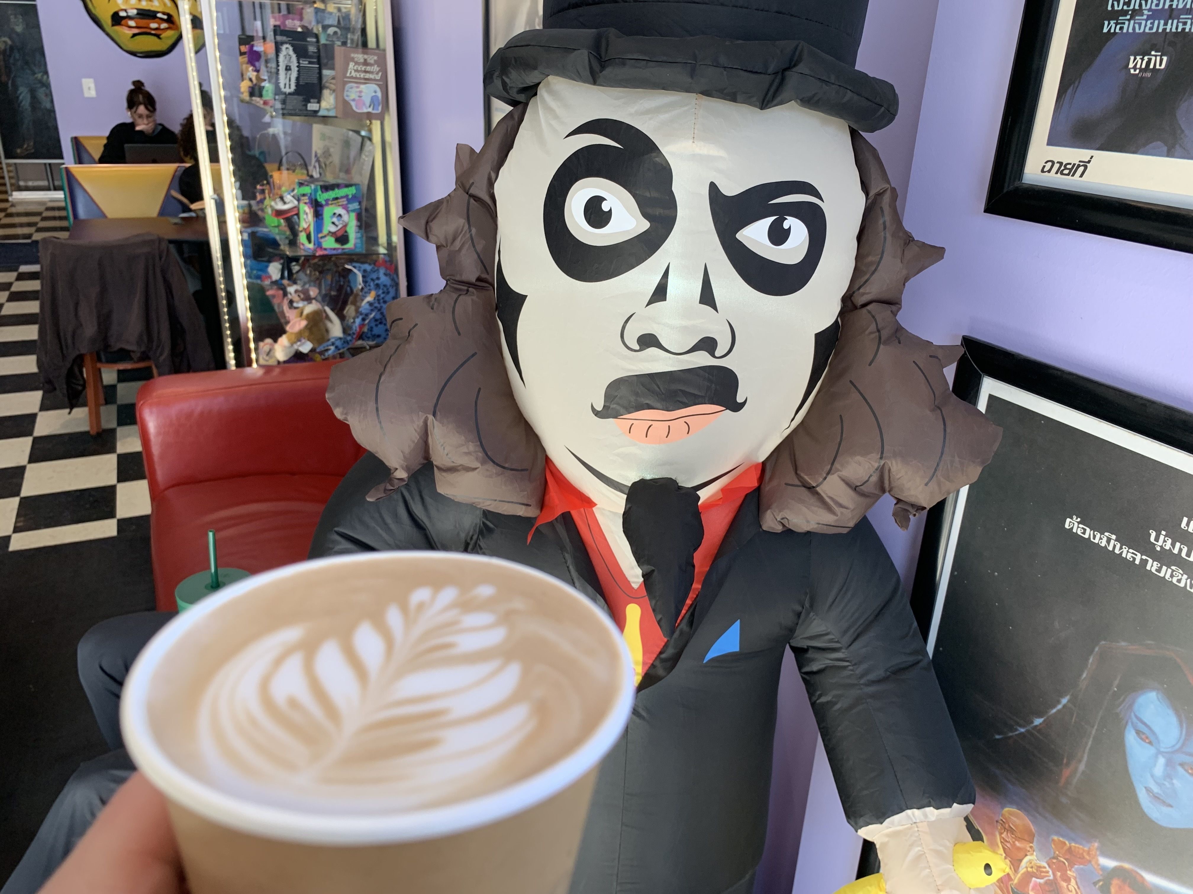 Photo of a cup of coffee next to a blow up doll. 